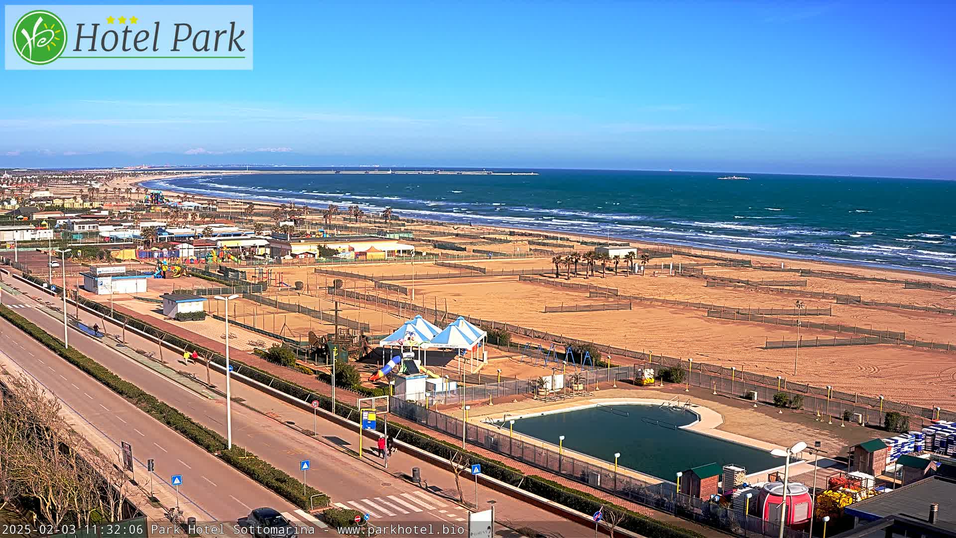 A beachside recreational area with a swimming pool, playground equipment, and volleyball courts is shown on a sunny day with wavy ocean waters in the background.