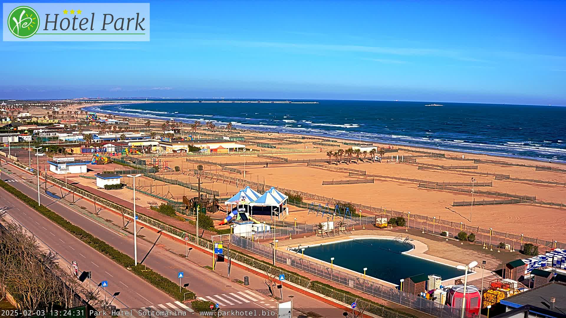 A wide shot of a beach on a sunny day shows a sandy beach with volleyball courts, a playground, a swimming pool, and a road leading to it.