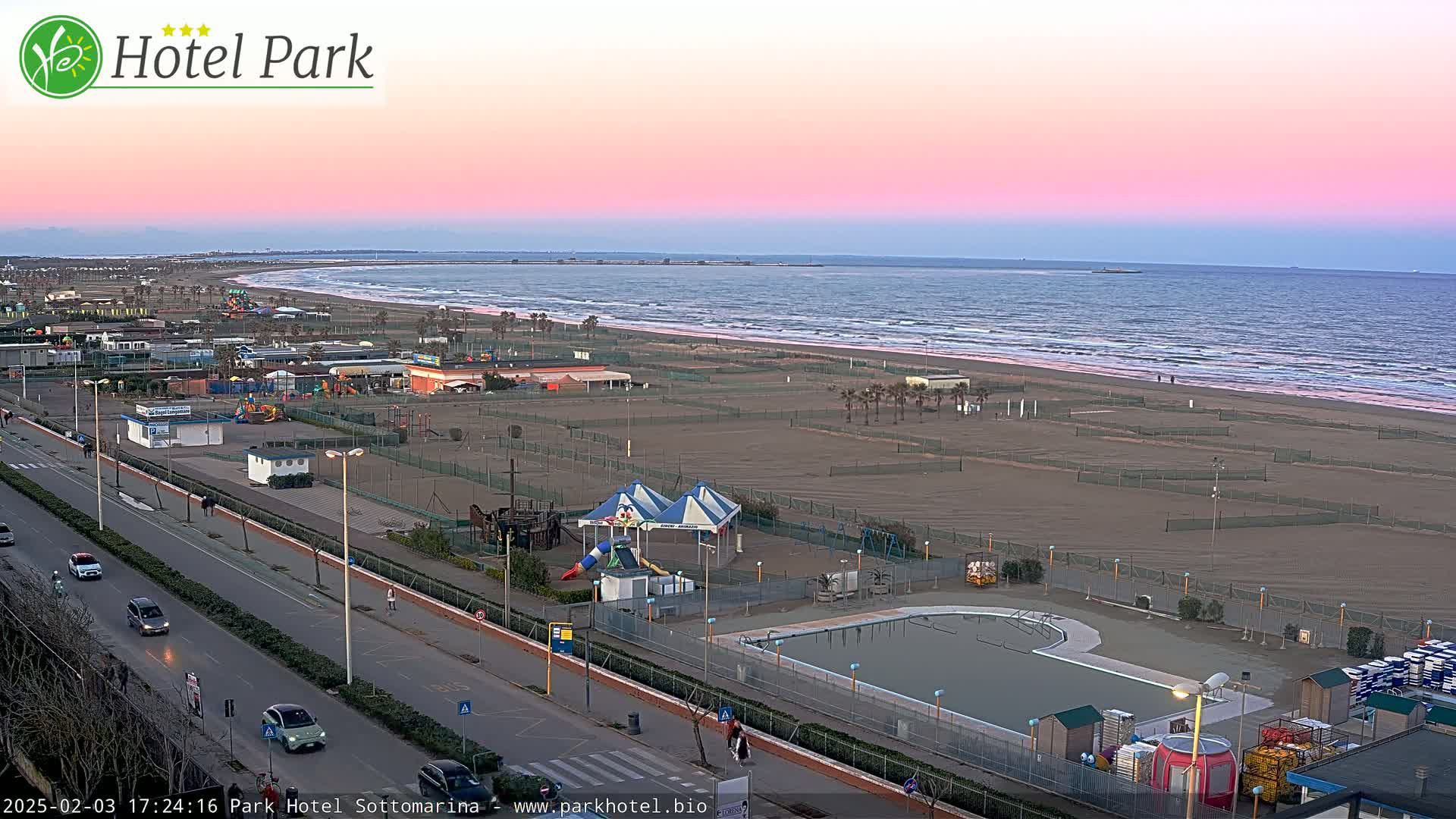 A beachside amusement area with a swimming pool, playground, and a calm ocean under a pinkish-purple sunset sky.