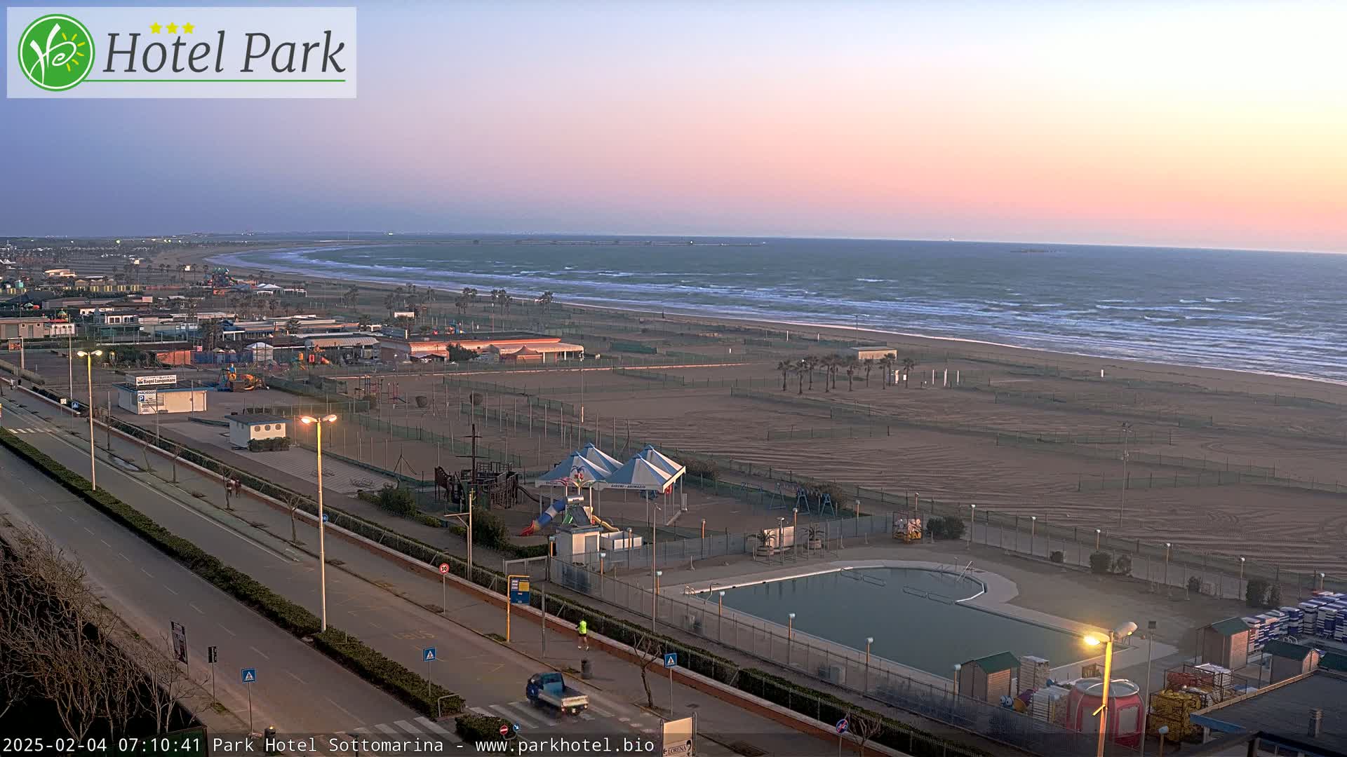 An expansive beach with a playground, swimming pool, and road is visible during a pink-hued sunrise, with somewhat choppy waves in the ocean.