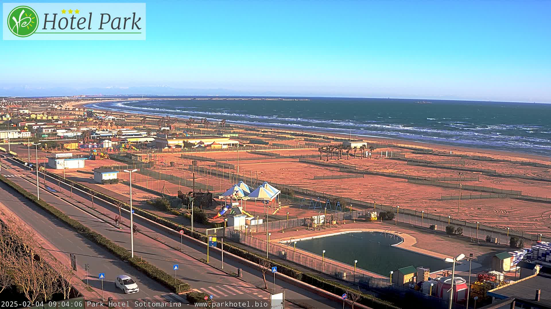A beachside amusement area with a pool and playground is visible, next to a road and a windy ocean with waves under a clear blue sky.