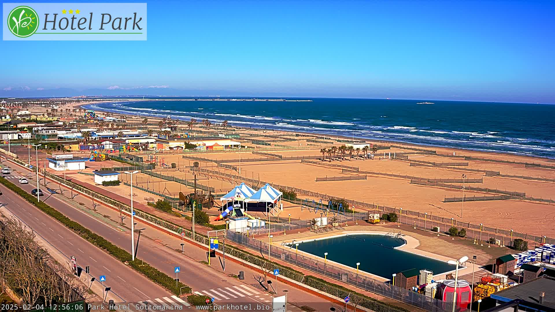 A beach with a playground, swimming pool, and a road leading to it on a sunny day with wavy ocean waters in the background.