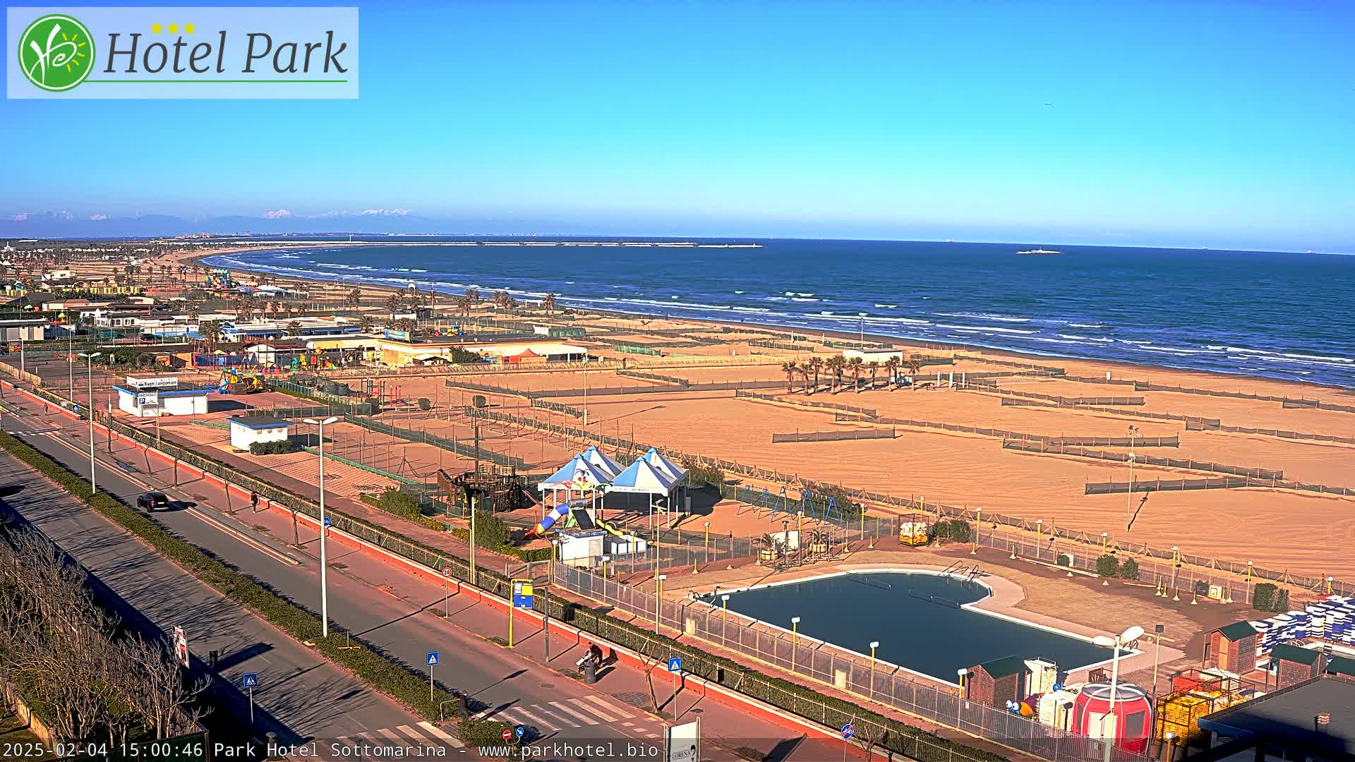 A beach with a swimming pool and playground is visible from a high vantage point on a sunny day, with mountains in the distance and a calm sea.