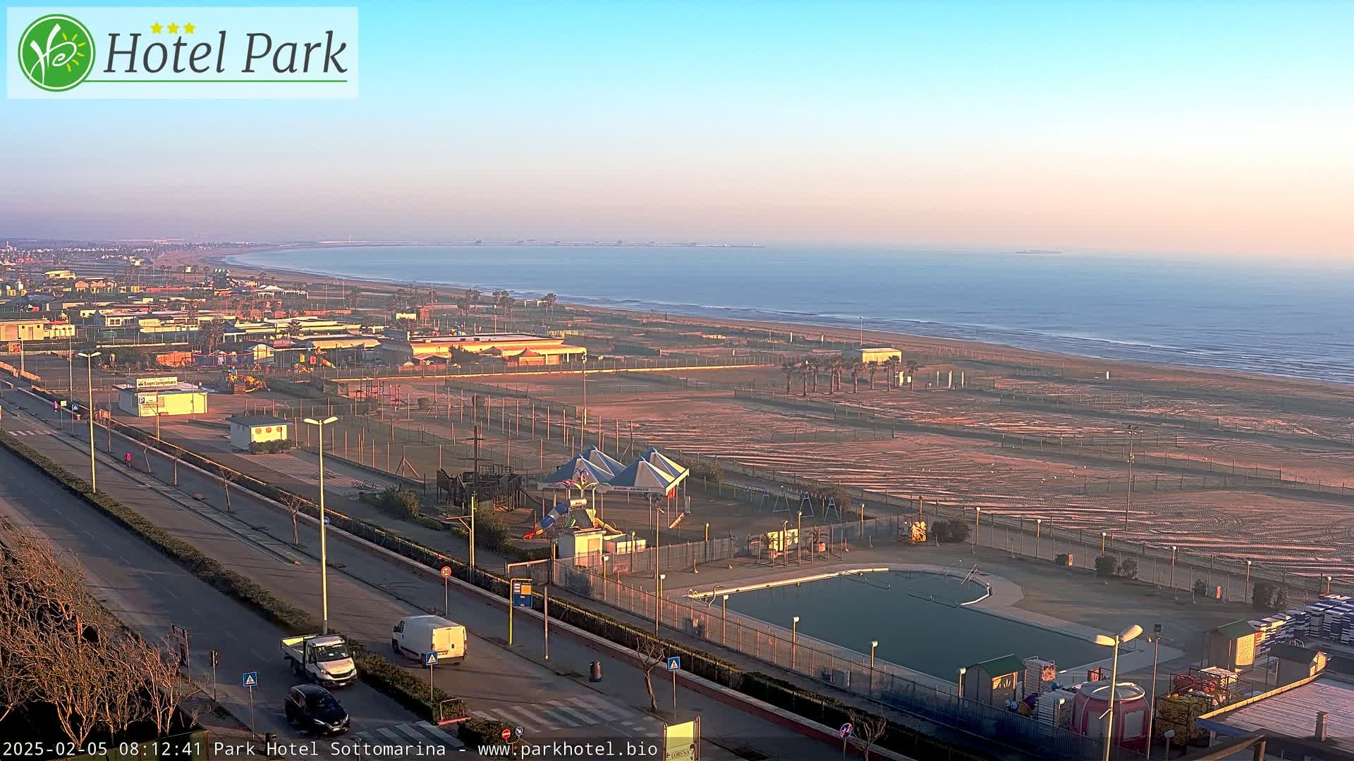 A coastal area with a beach, a swimming pool, and a playground is seen at sunrise under a clear sky.