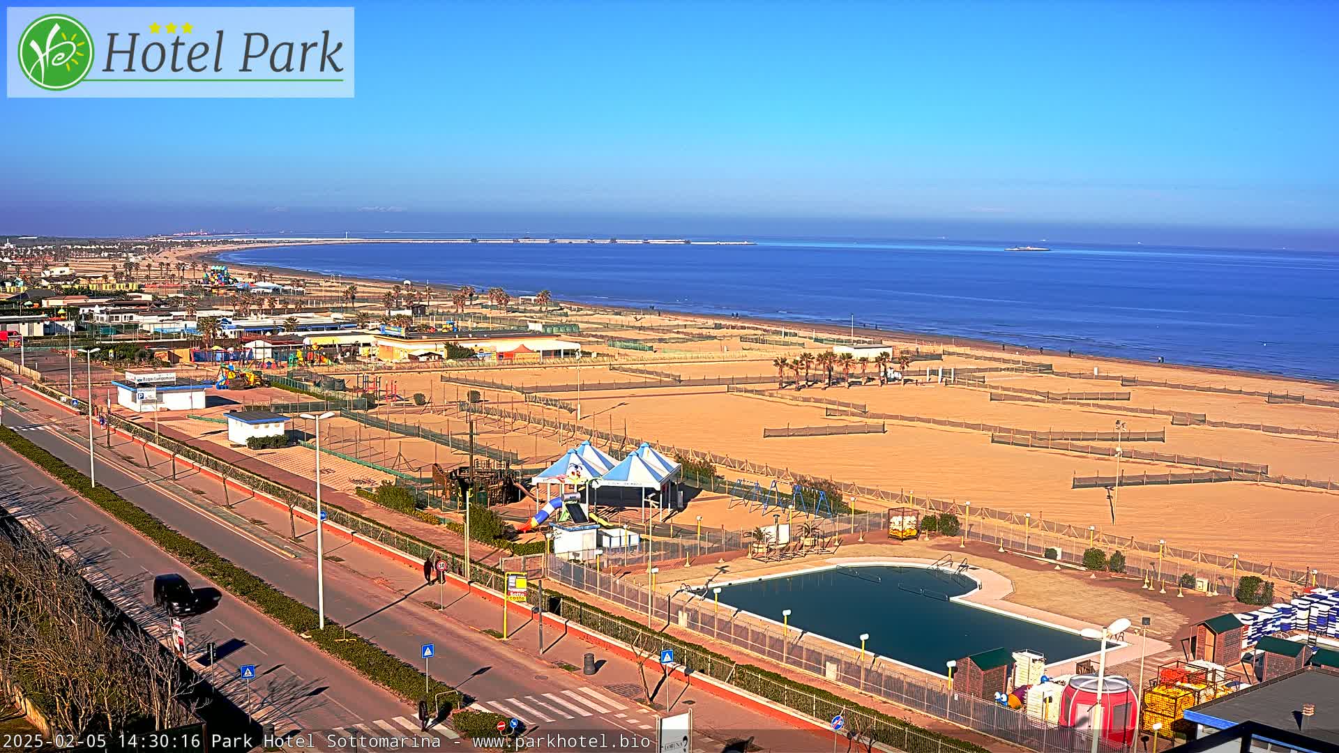A long sandy beach with volleyball courts, a swimming pool, playground equipment, and a road leading to it on a clear sunny day.