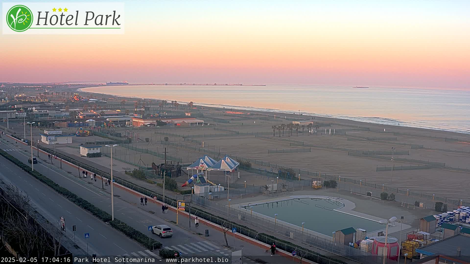 A long sandy beach at sunset, with a swimming pool, playground equipment, and a road running parallel to the shore.