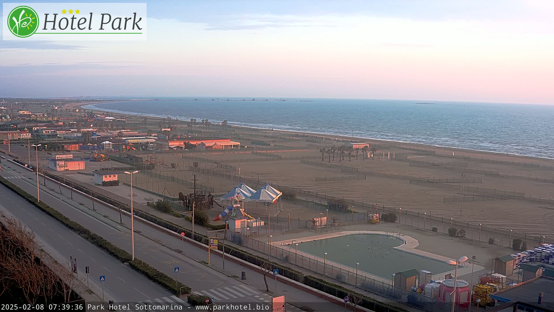 A long sandy beach stretches along a calm ocean under a pale pink sunrise, with a road and playground visible closer to the camera.