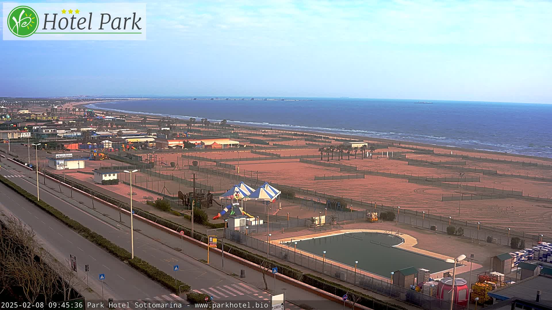 A beachside resort area with a swimming pool, playground, and numerous sand courts is visible on a partly cloudy day, overlooking a calm sea.