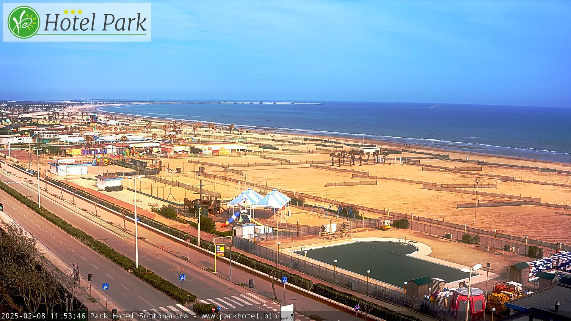 A beachside amusement area with a swimming pool, playground equipment, and volleyball courts is shown, with a clear, sunny sky and calm ocean visible in the background.