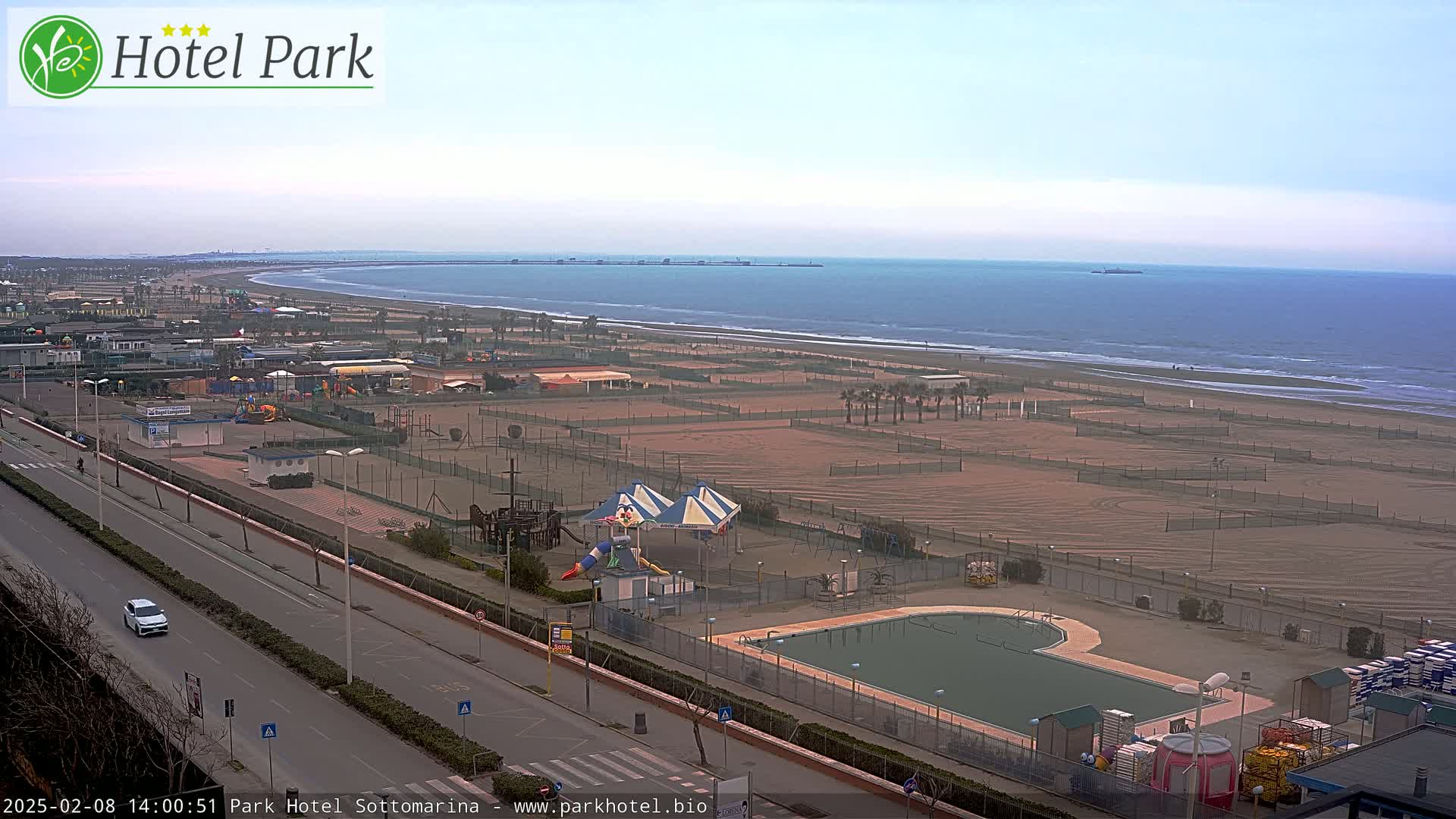 A mostly clear day overlooking a beach with volleyball courts, a playground, and a swimming pool, all situated next to a road.