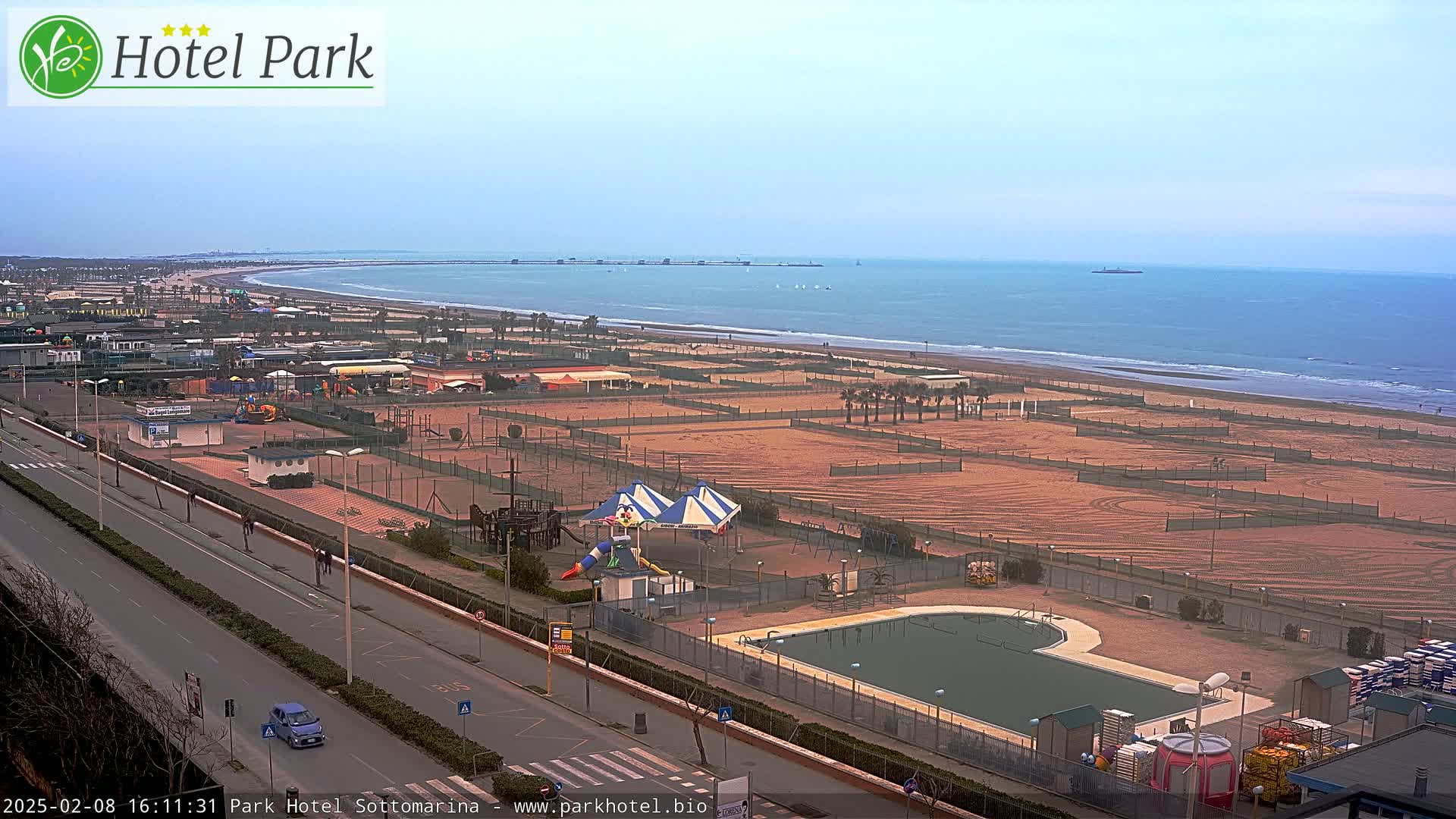 An aerial view shows a beach with volleyball courts and a playground next to a roadway, a swimming pool, and a calm ocean under a mostly clear sky.