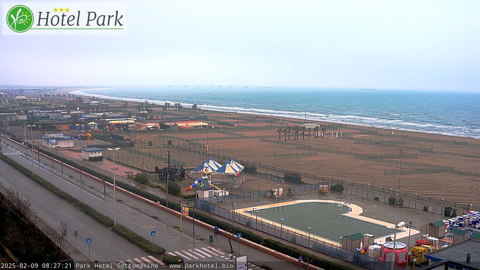 An overcast day reveals a beach with a mostly empty sandy expanse, bordered by a road, playground equipment, and a swimming pool, all overlooking a wavy ocean.