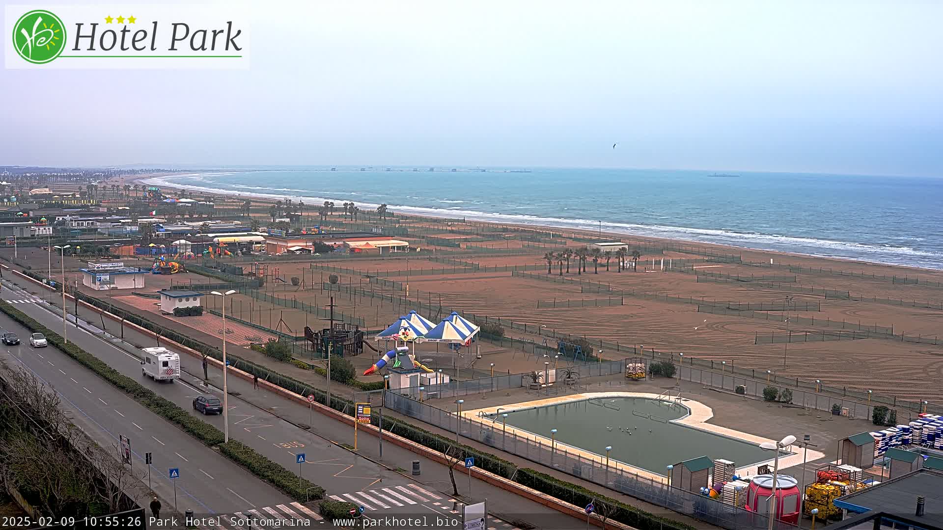 An overcast day at a beachside resort shows a wide sandy beach, a swimming pool, playground equipment, and a roadway with vehicles.