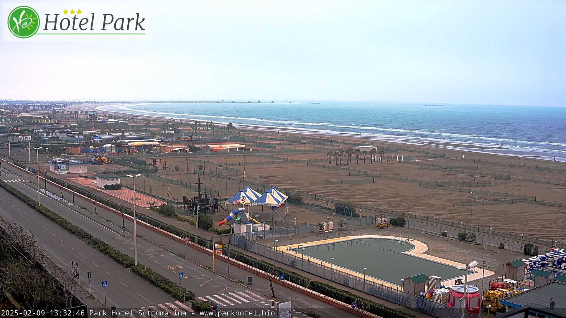 An aerial view shows a beach with calm waves, a playground, a swimming pool, and a road next to the ocean on an overcast day.