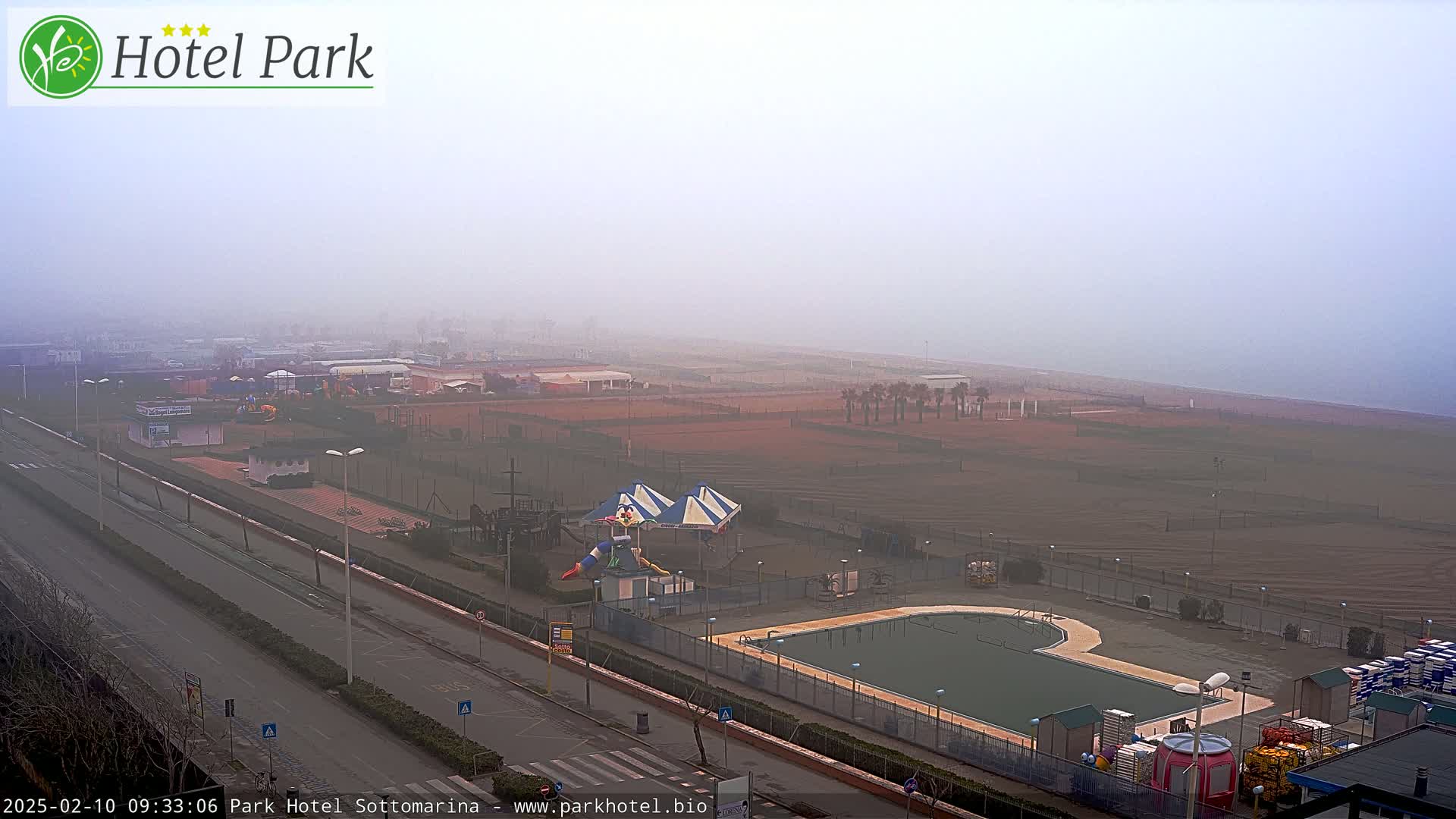 A foggy view of a beachside resort area, featuring a swimming pool, playground equipment, and a roadway.