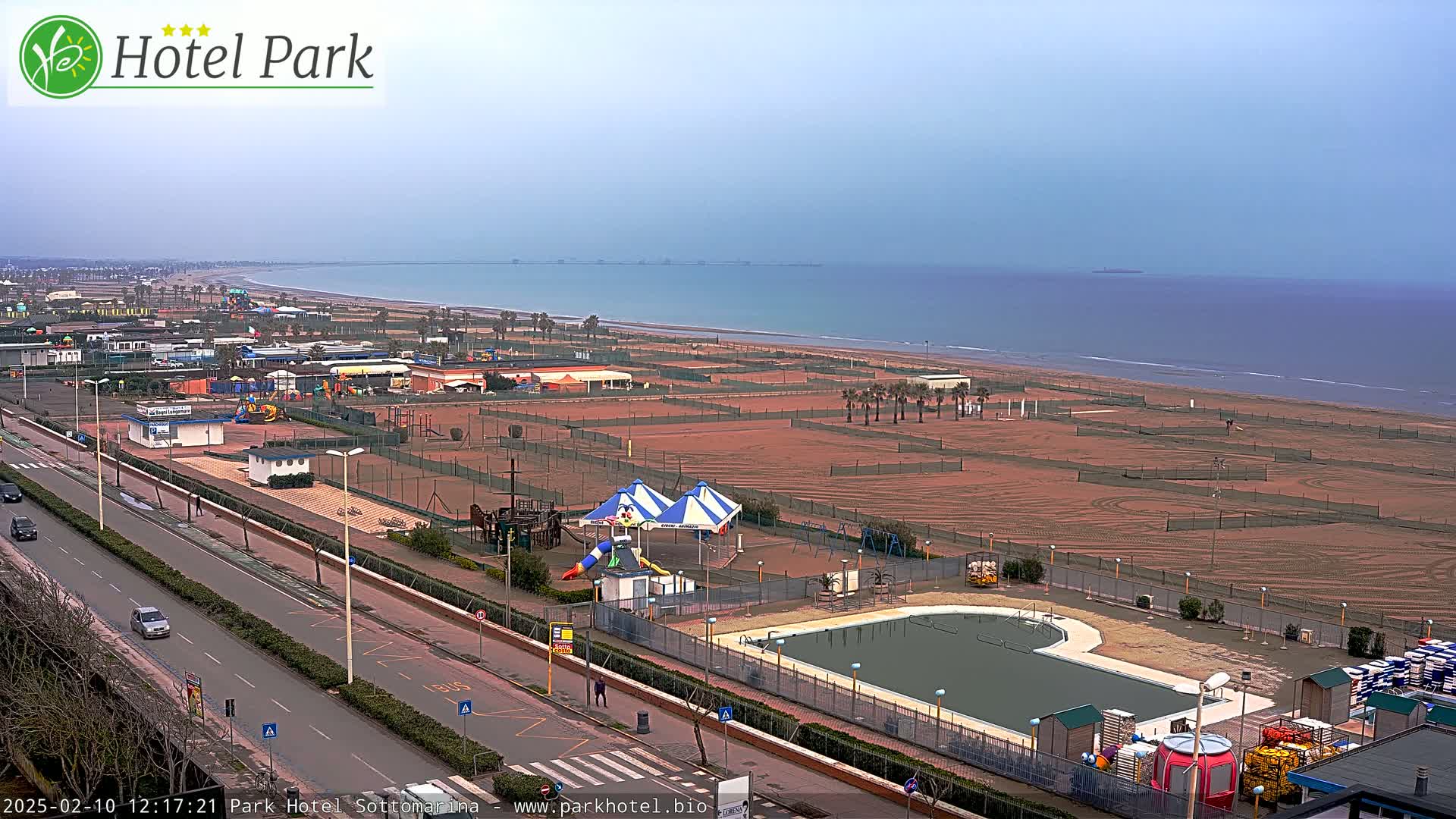 An overcast day reveals a beach with a playground, swimming pool, and tennis courts adjacent to a road, all overlooking a calm sea.