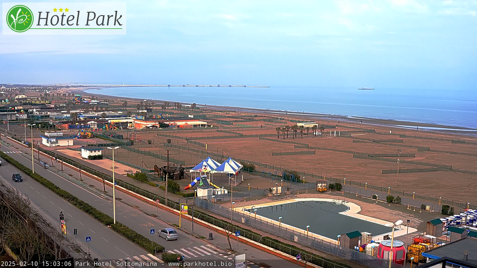 A beachfront resort area with a swimming pool, playground equipment, and tennis courts under a mostly clear sky.