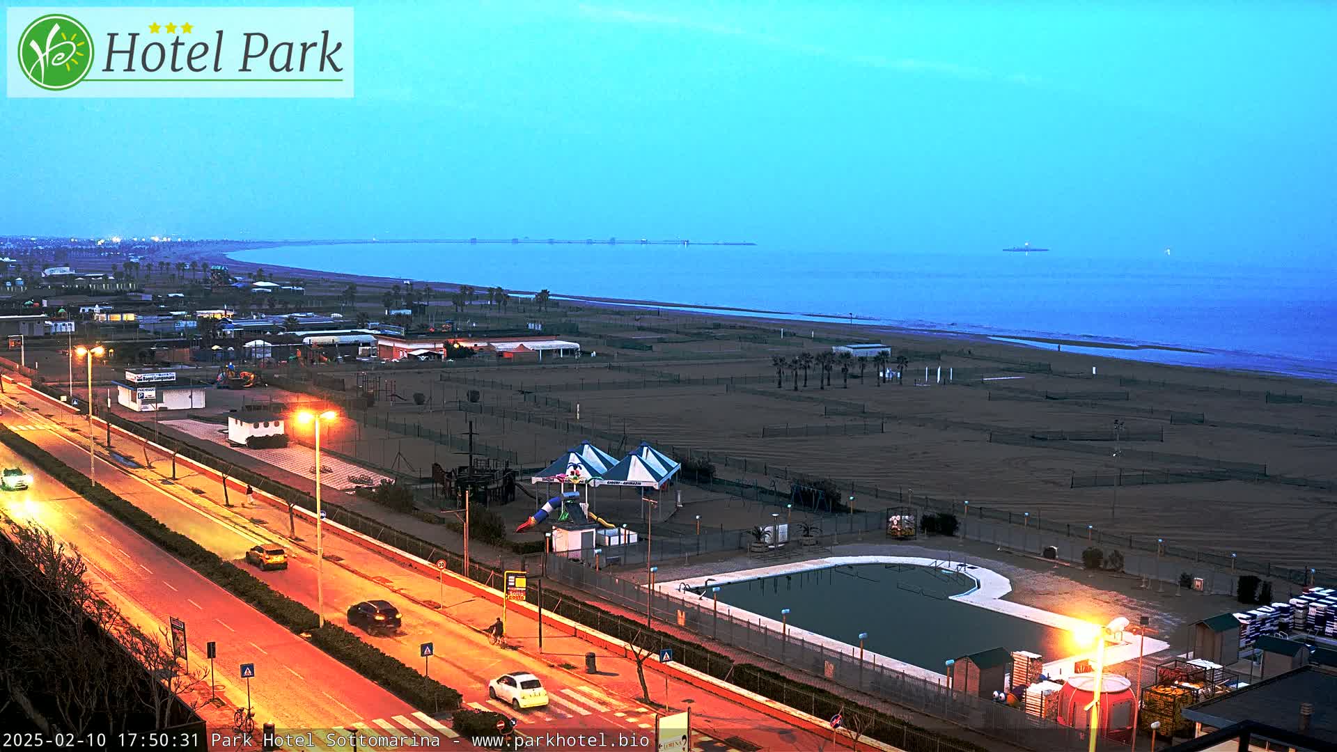 A nighttime, coastal view shows a beach, a road with cars, a swimming pool, and playground equipment under a twilight sky.