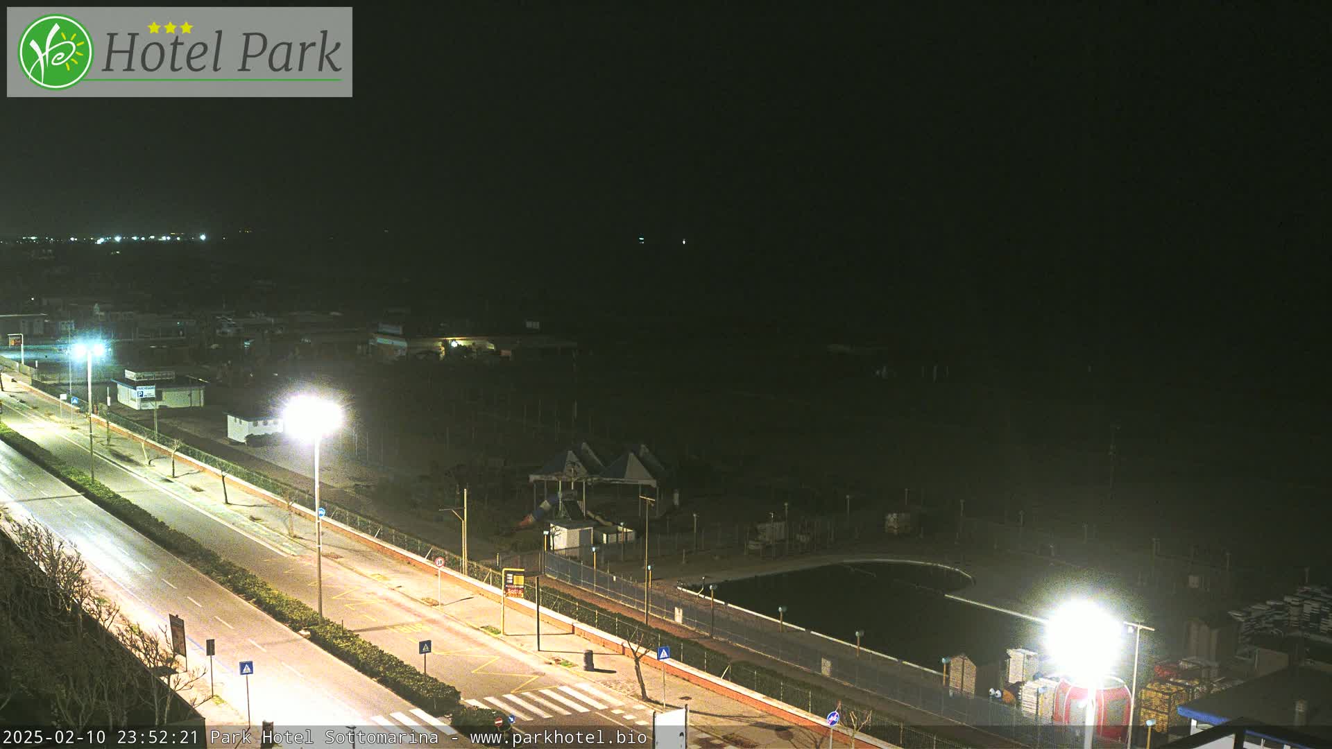 A nighttime view of a mostly empty road next to a park with a pool, playground equipment, and some buildings under a clear, dark sky.
