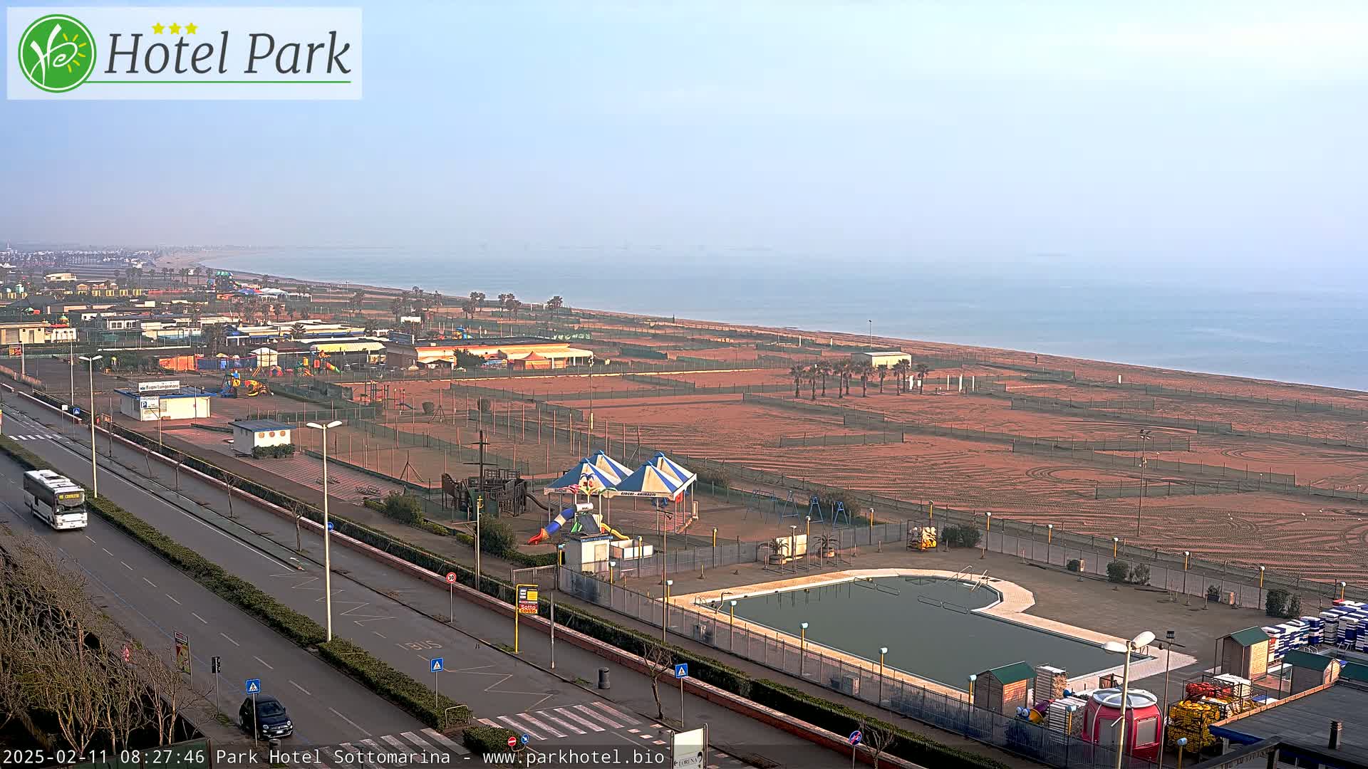 An aerial view shows a beachfront resort with a swimming pool, playground, and sandy beach on a hazy day.