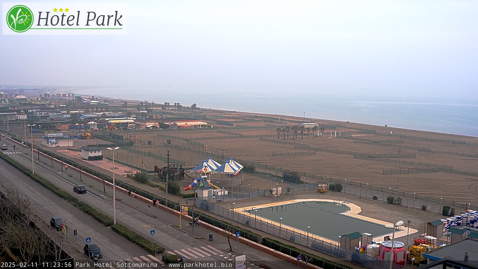 A hazy, overcast day reveals a beachfront area with a swimming pool, playground, and sandy beach extending to a calm sea.