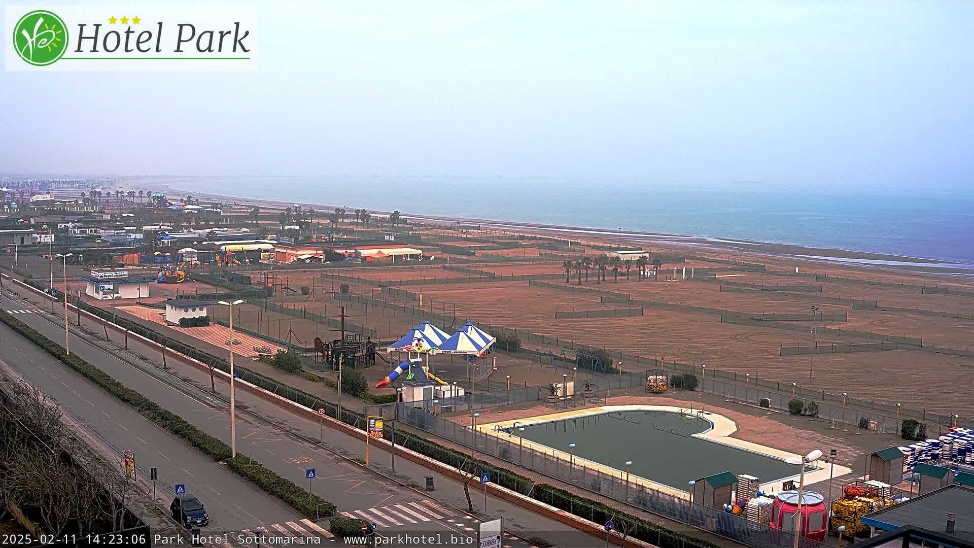 A hazy coastal view shows a resort area with a swimming pool, playground, and tennis courts adjacent to a wide sandy beach and calm sea.