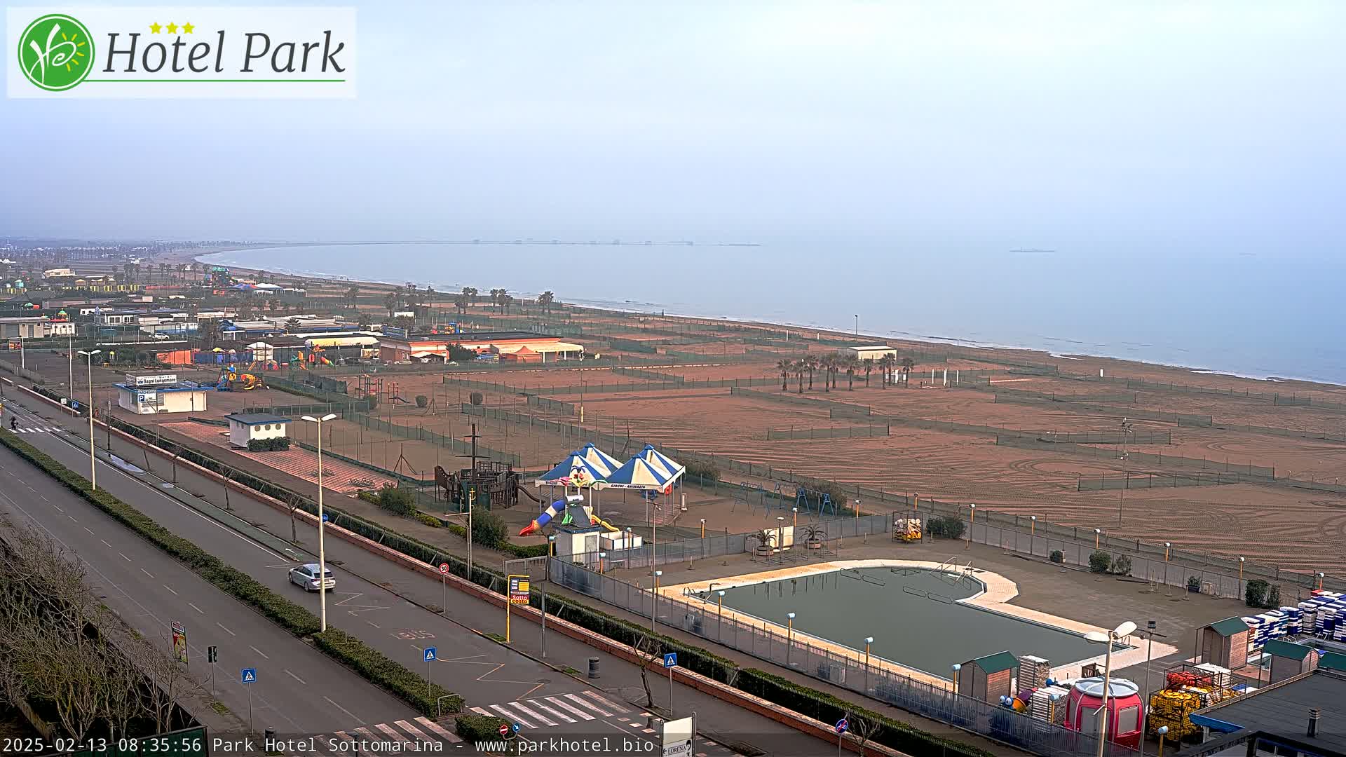 An aerial view of a beachfront resort area with a playground, swimming pool, and empty sandy beach on an overcast day.