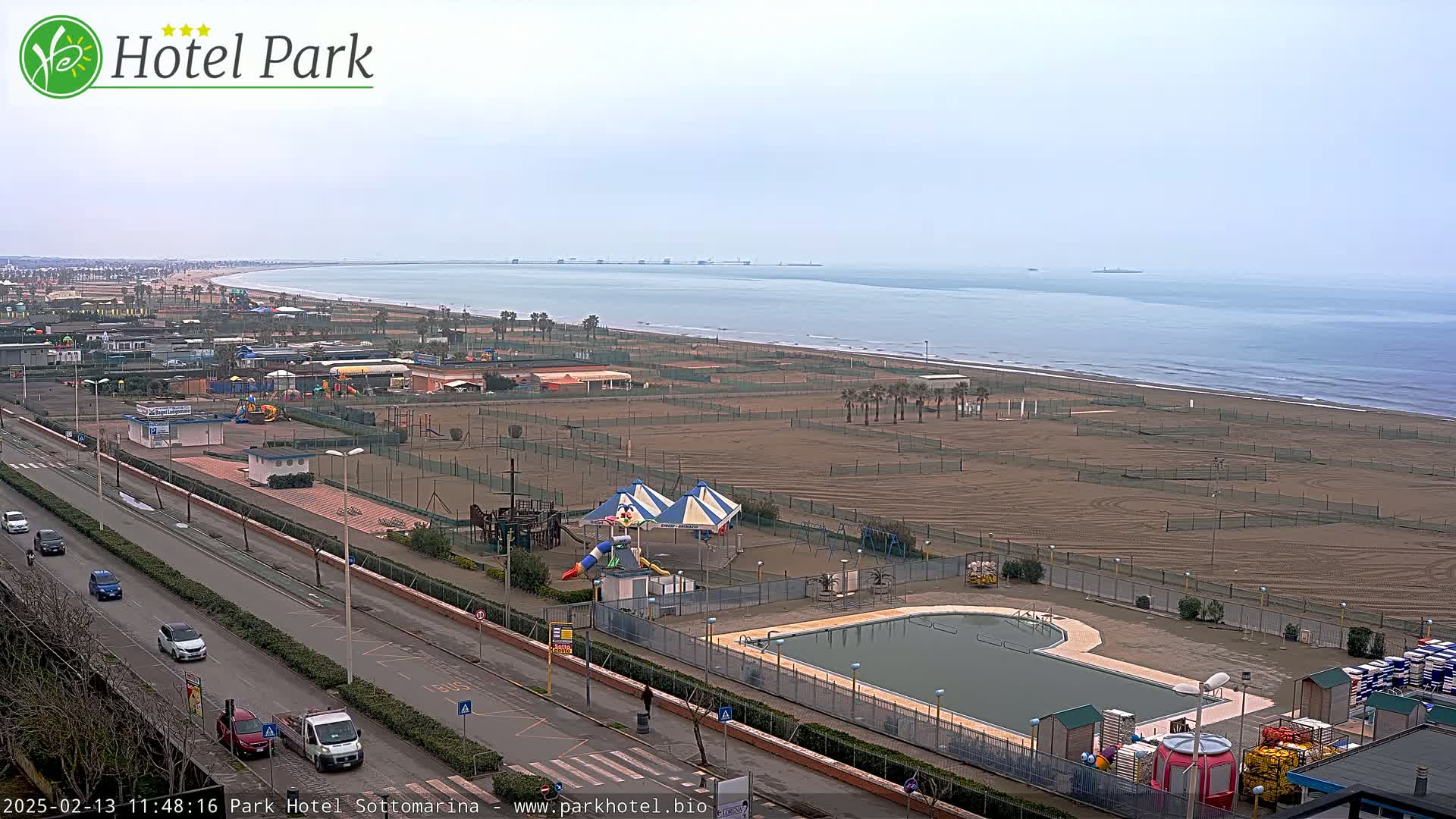 An overcast day shows a beach with a playground, swimming pool, and a road with cars parked along the side.