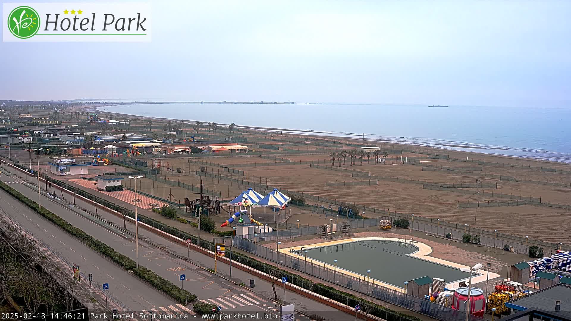 An overcast day reveals a beach with a playground, swimming pool, and roadway next to it, all overlooking a calm sea.