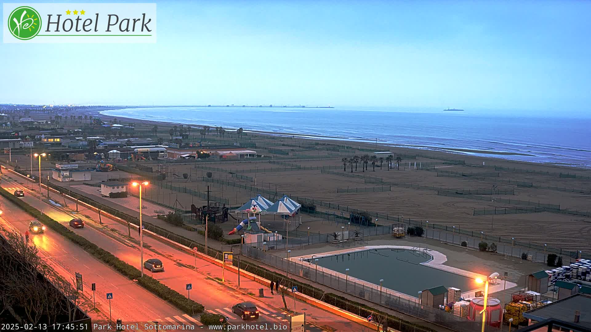 An aerial view of a beach at dusk, showing a road with cars, a playground, a swimming pool, and a calm ocean.