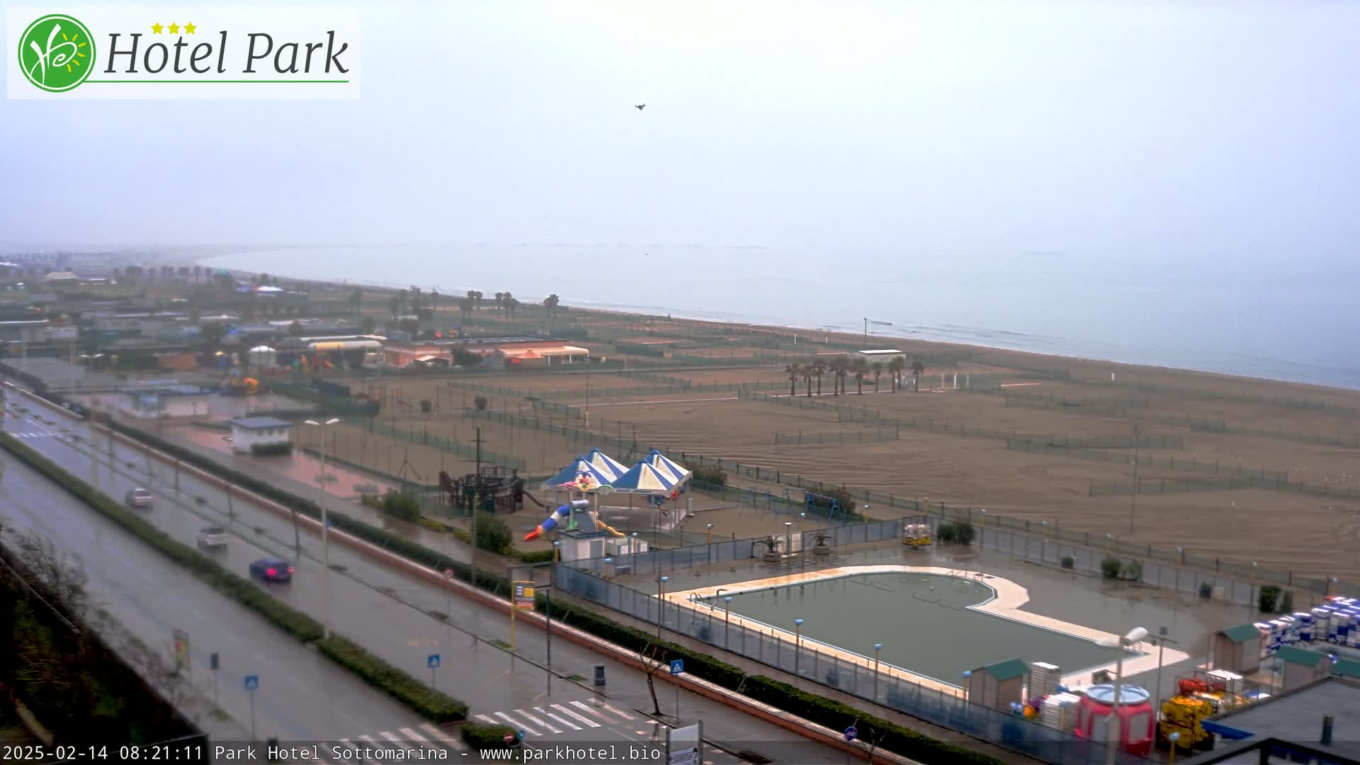 A rain-soaked coastal area with a flooded pool and playground near a road and beach under an overcast sky.