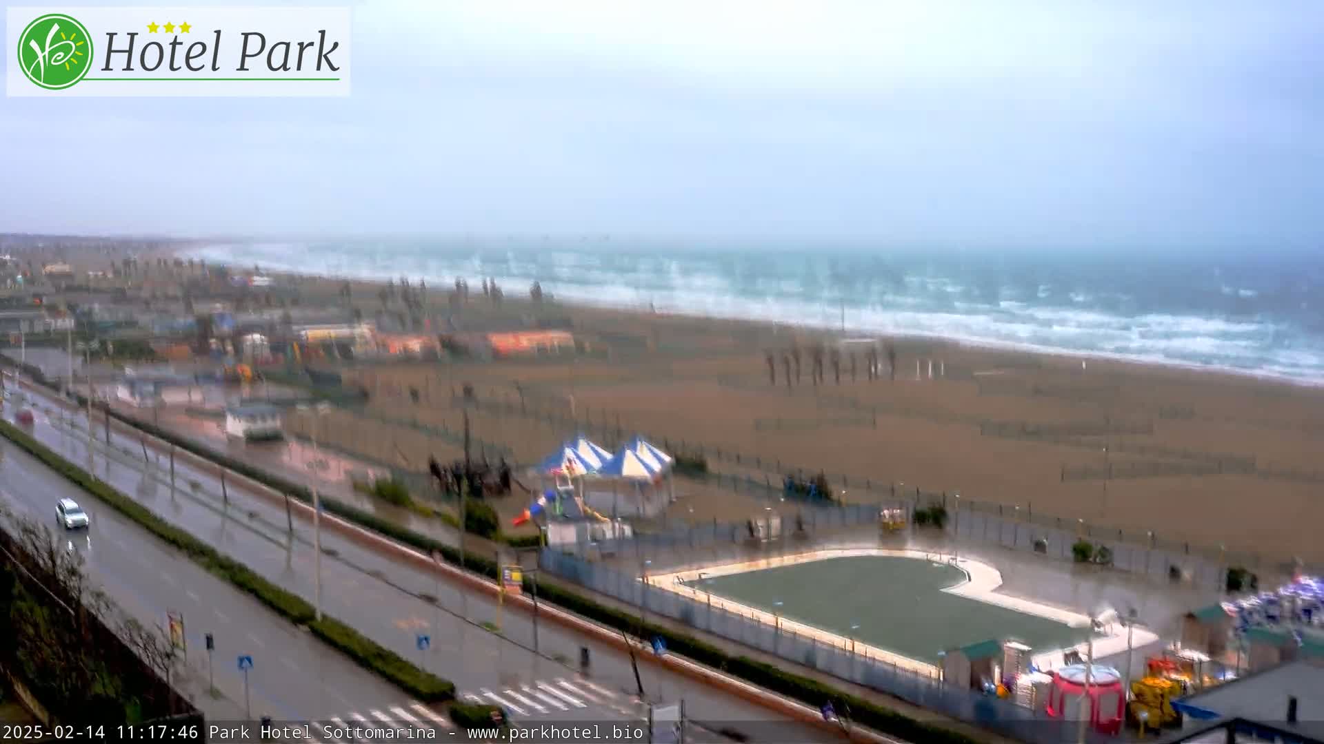 A rain-swept beach scene shows choppy waves, a wet roadway, and a partially flooded beachside area with playground equipment near a pool.
