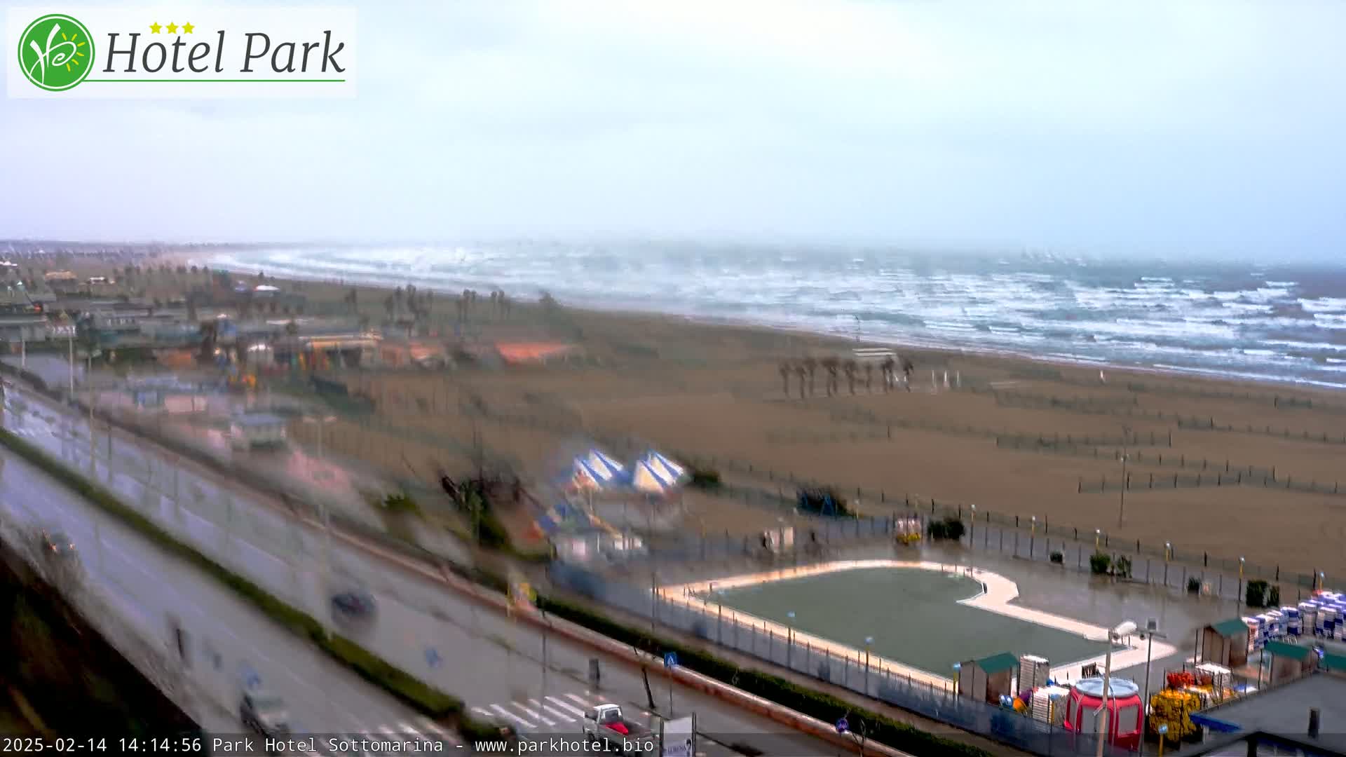 A rain-swept beach scene shows a wide, sandy beach with choppy waves, a road with moving vehicles, and a swimming pool area with playground equipment nearby.