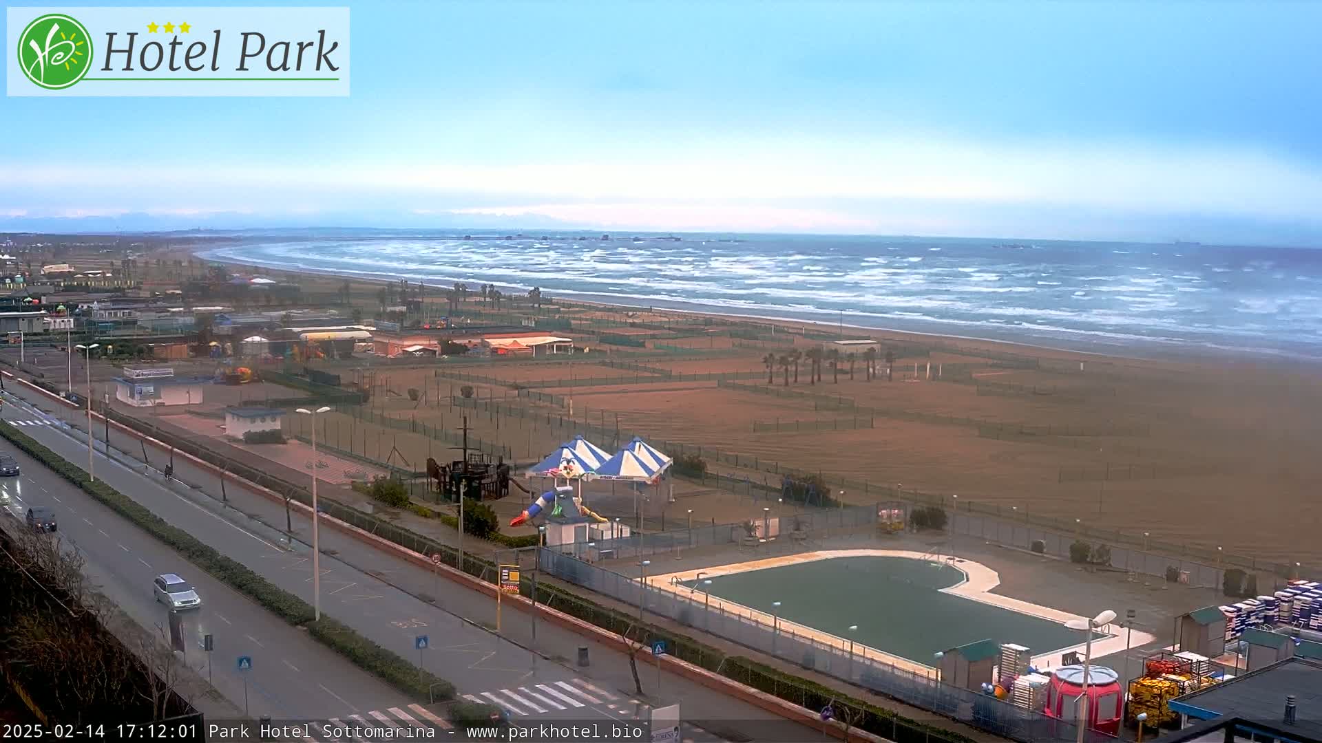 A beach with a playground, swimming pool, and a road is seen under a mostly cloudy sky with a windy ocean in the background.