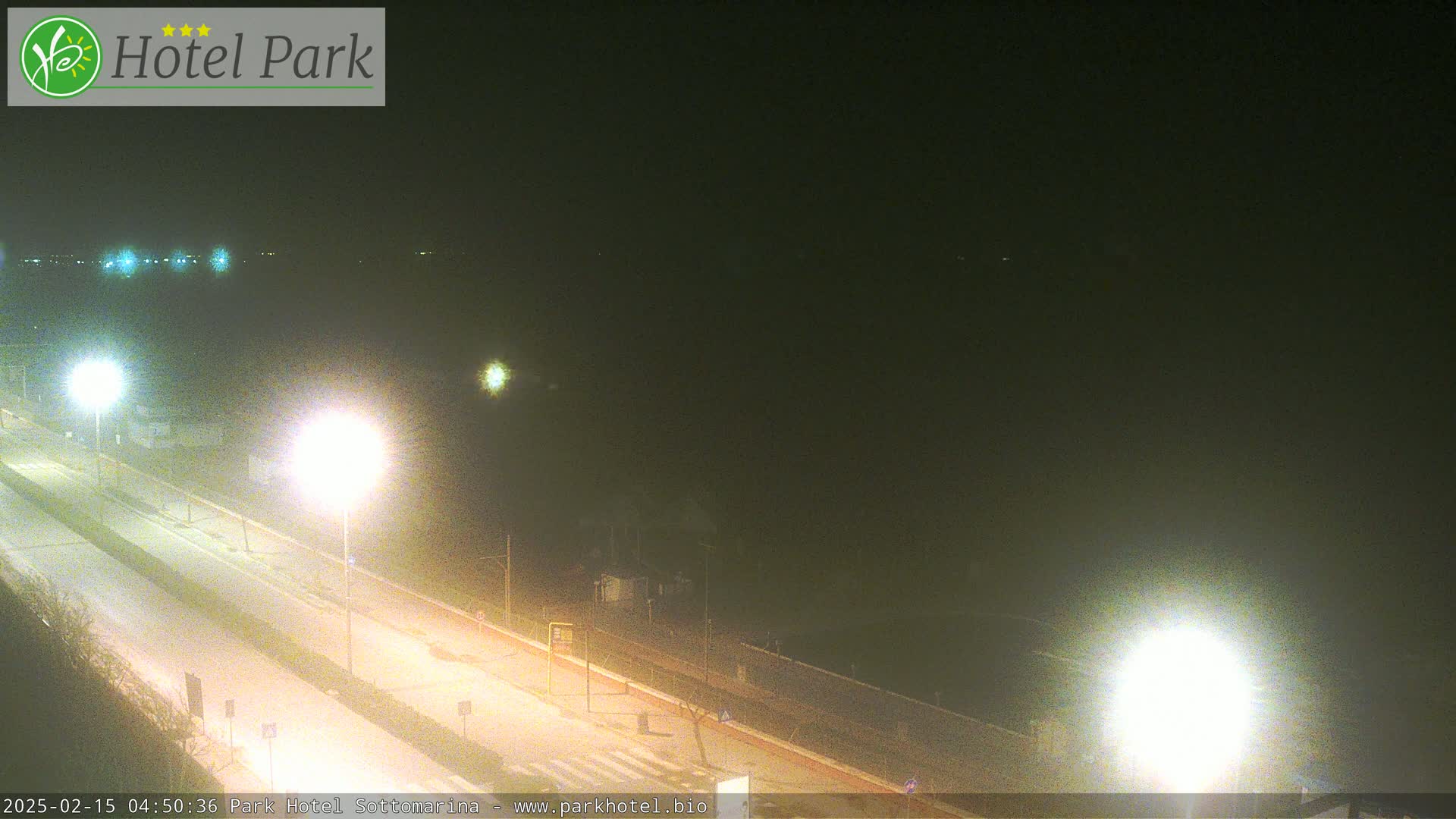 A nighttime, hazy view of a mostly empty road beside a body of water, illuminated by streetlights.