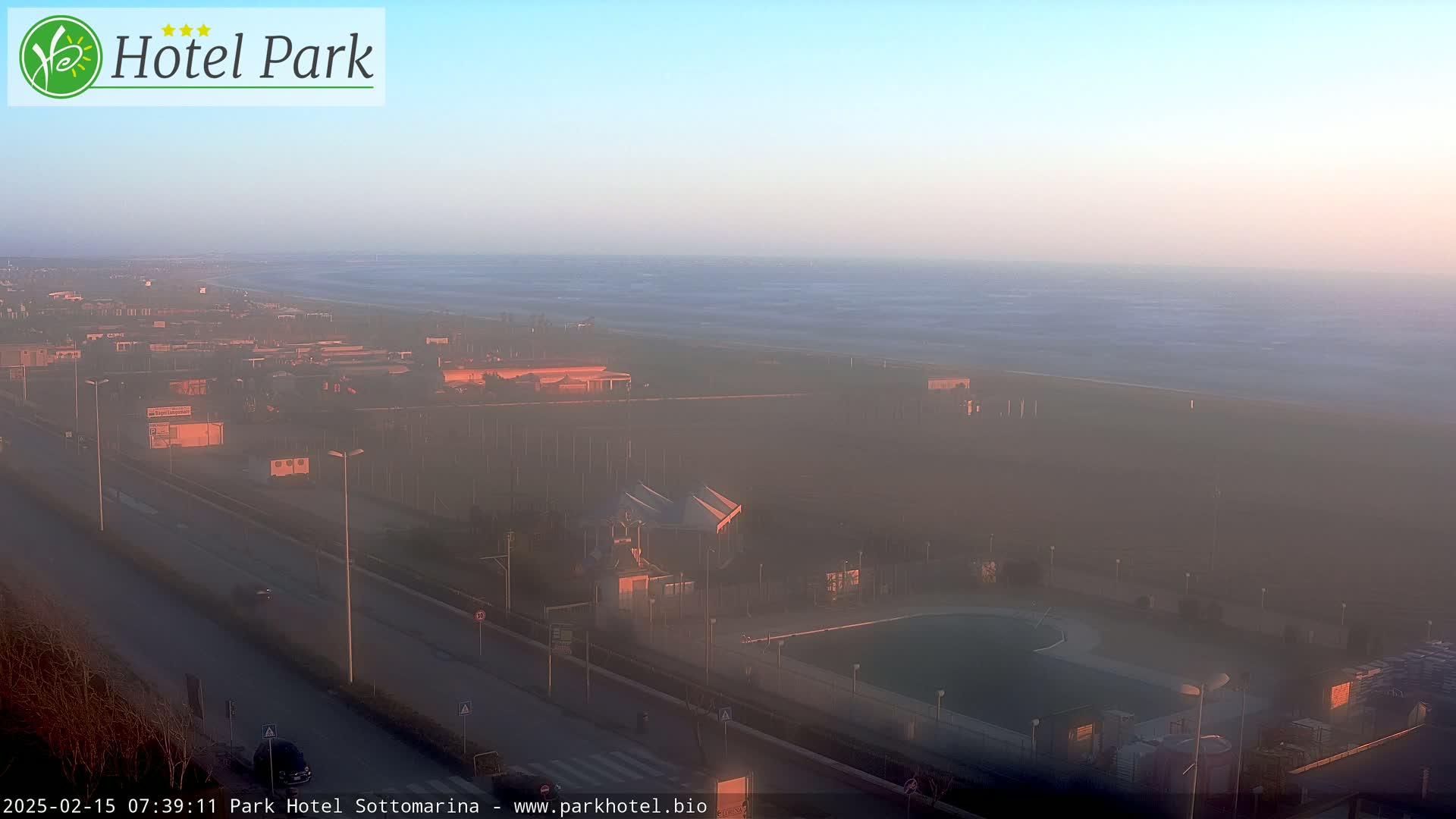 A hazy sunrise illuminates a coastal resort area with a beach, pool, and buildings, under a mostly clear sky.