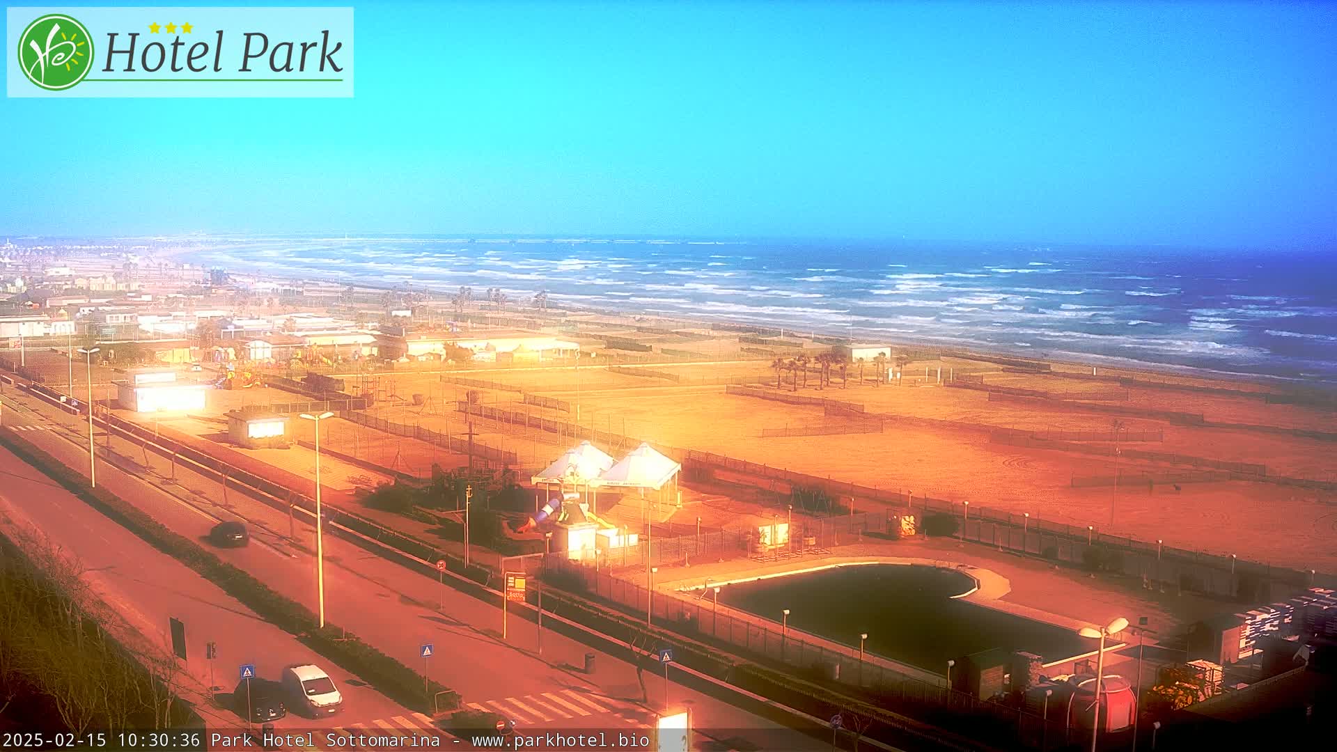 A beachside area with a pool, playground equipment, and a road, under a clear, bright sky, with a wavy ocean in the background.