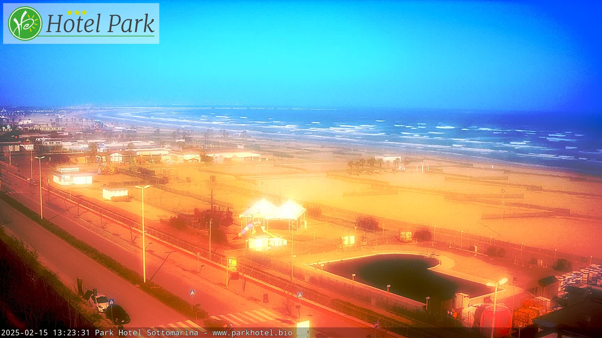 An aerial view shows a beach with calm waves under a clear, bright blue sky, featuring a sandy area with buildings, a swimming pool, and playground equipment.