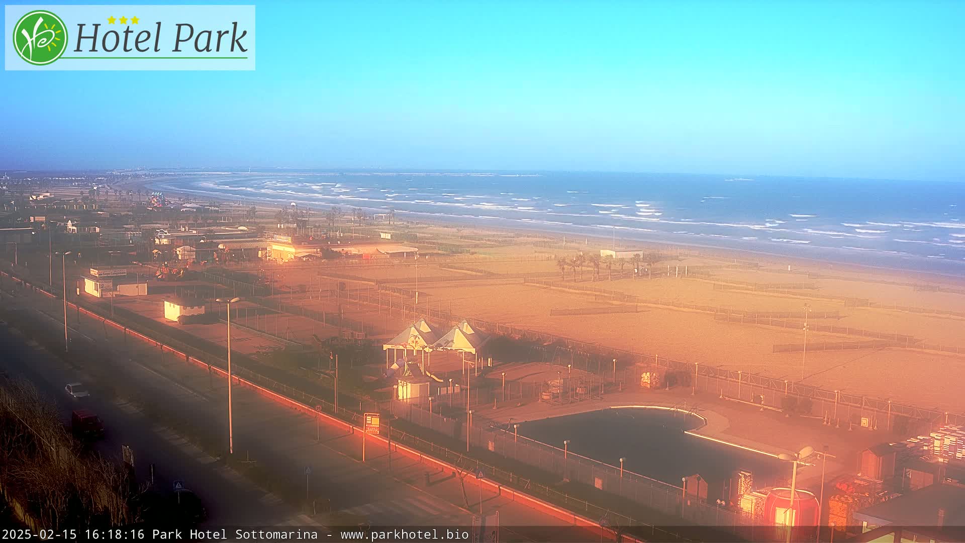 An expansive beach with gently rolling waves under a clear, bright sky is viewed from above, showing a beachfront area with a swimming pool and other recreational facilities.