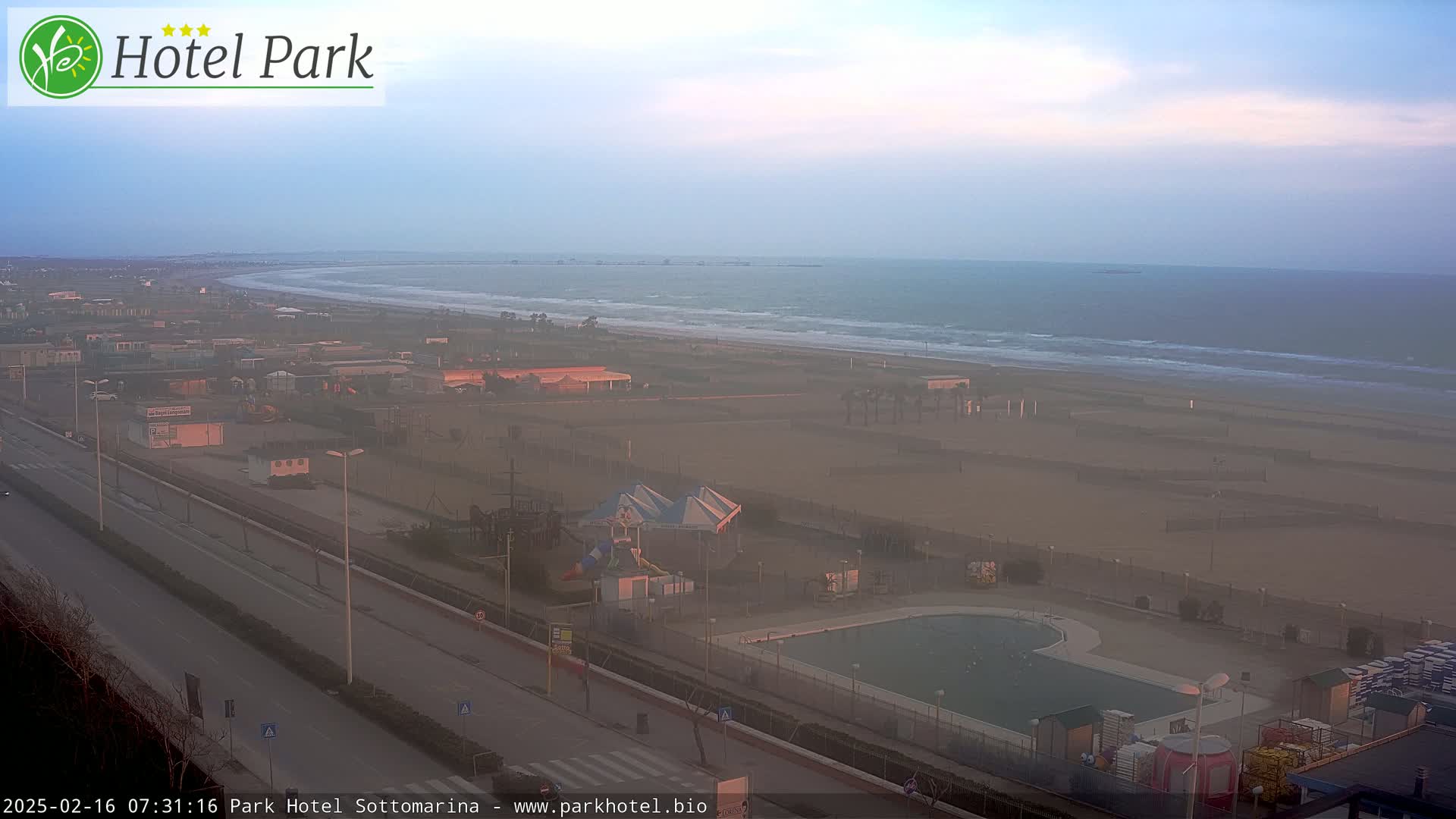 A hazy sunrise illuminates a beach with a swimming pool, playground, and road visible from an elevated vantage point.