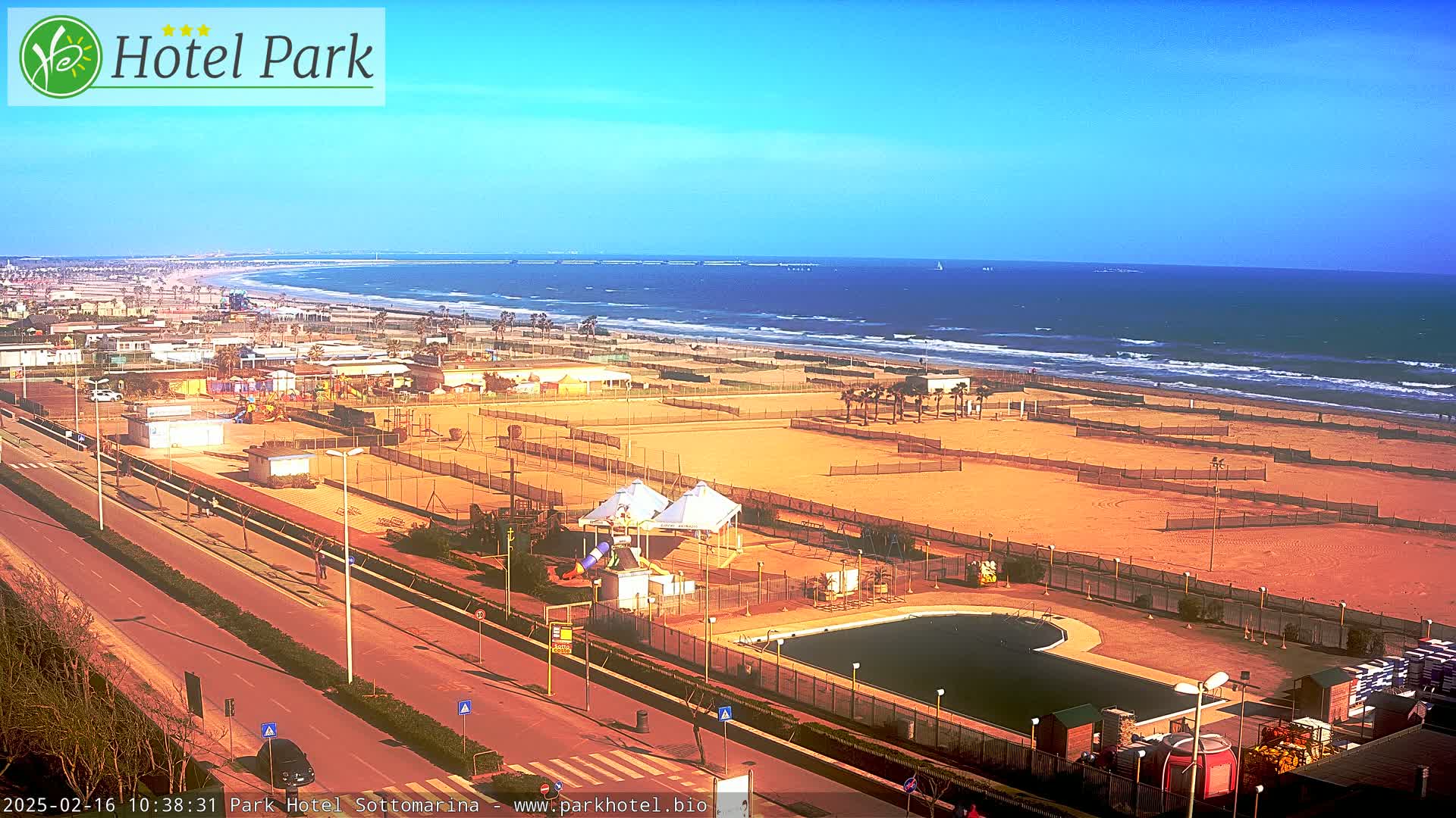 A sunny day reveals a beachside resort area with a pool, playground equipment, and tennis courts, all adjacent to a sandy beach and ocean with small waves.