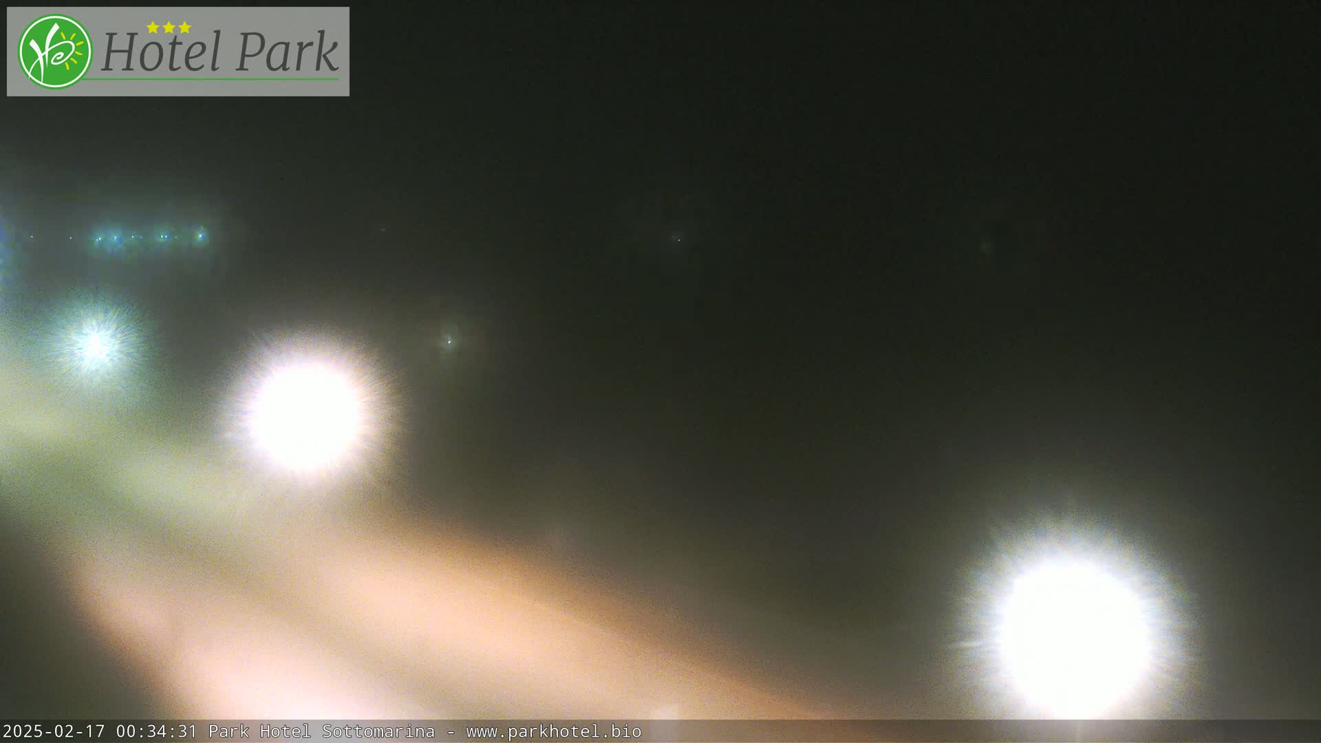 A nighttime, foggy scene shows the bright headlights of several vehicles on a road.