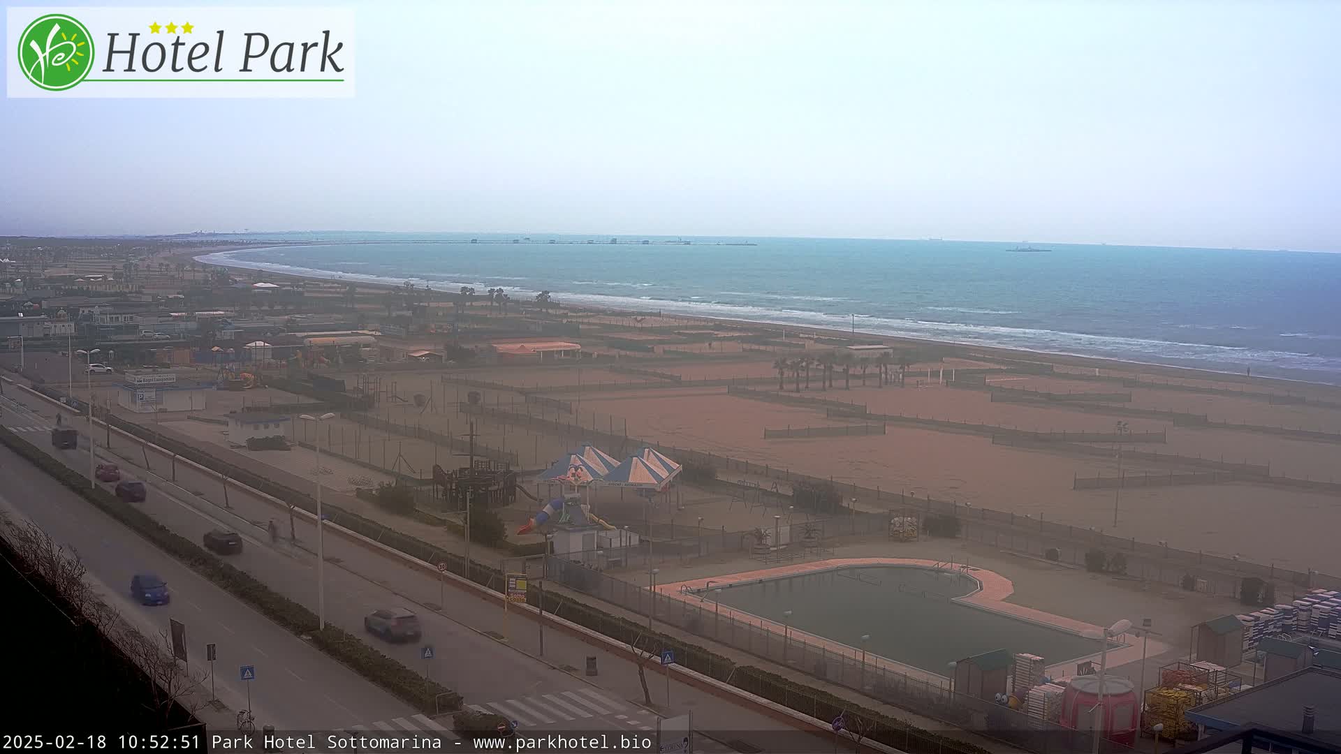 An overcast day reveals a beach with a swimming pool and playground near a road, all overlooking a calm ocean.
