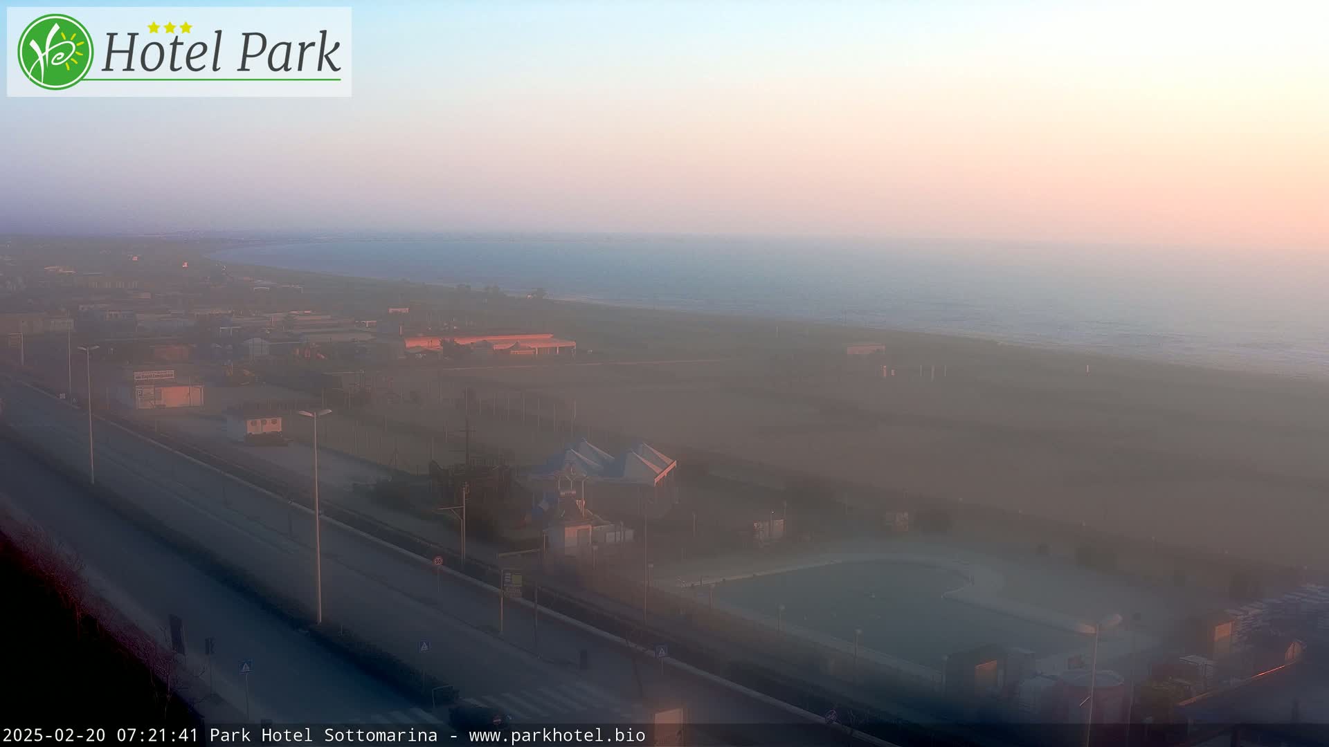 A hazy sunrise illuminates a coastal scene featuring a highway, buildings, a beach, and the ocean.