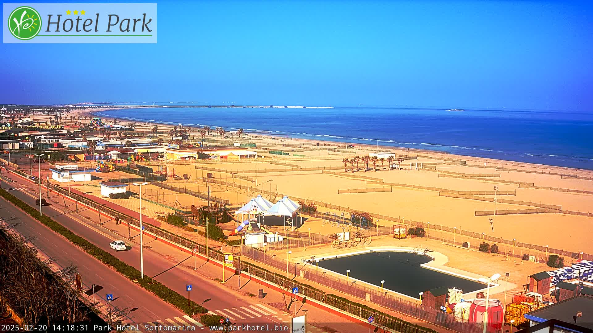 A beachside resort area with a swimming pool and playground is visible under a clear blue sky, with a calm ocean in the background.