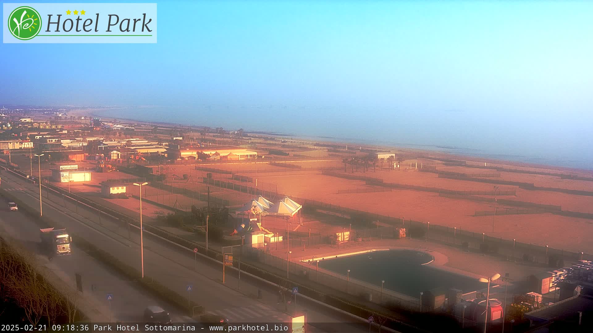 An aerial view shows a beach, a swimming pool, playground equipment, and a road, all bathed in the warm light of a hazy sunrise.