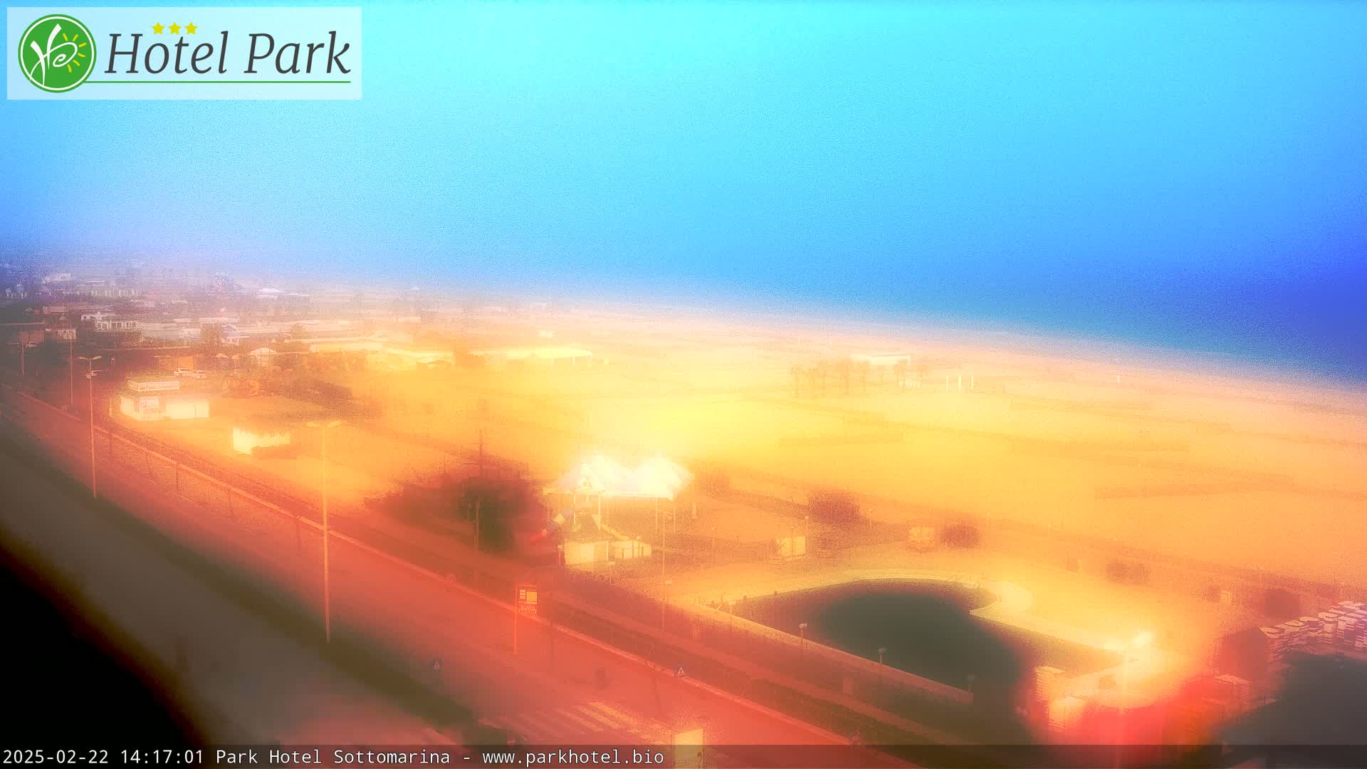 An elevated view shows a hazy, sunlit beach and surrounding area, with a pool and road visible in the foreground.