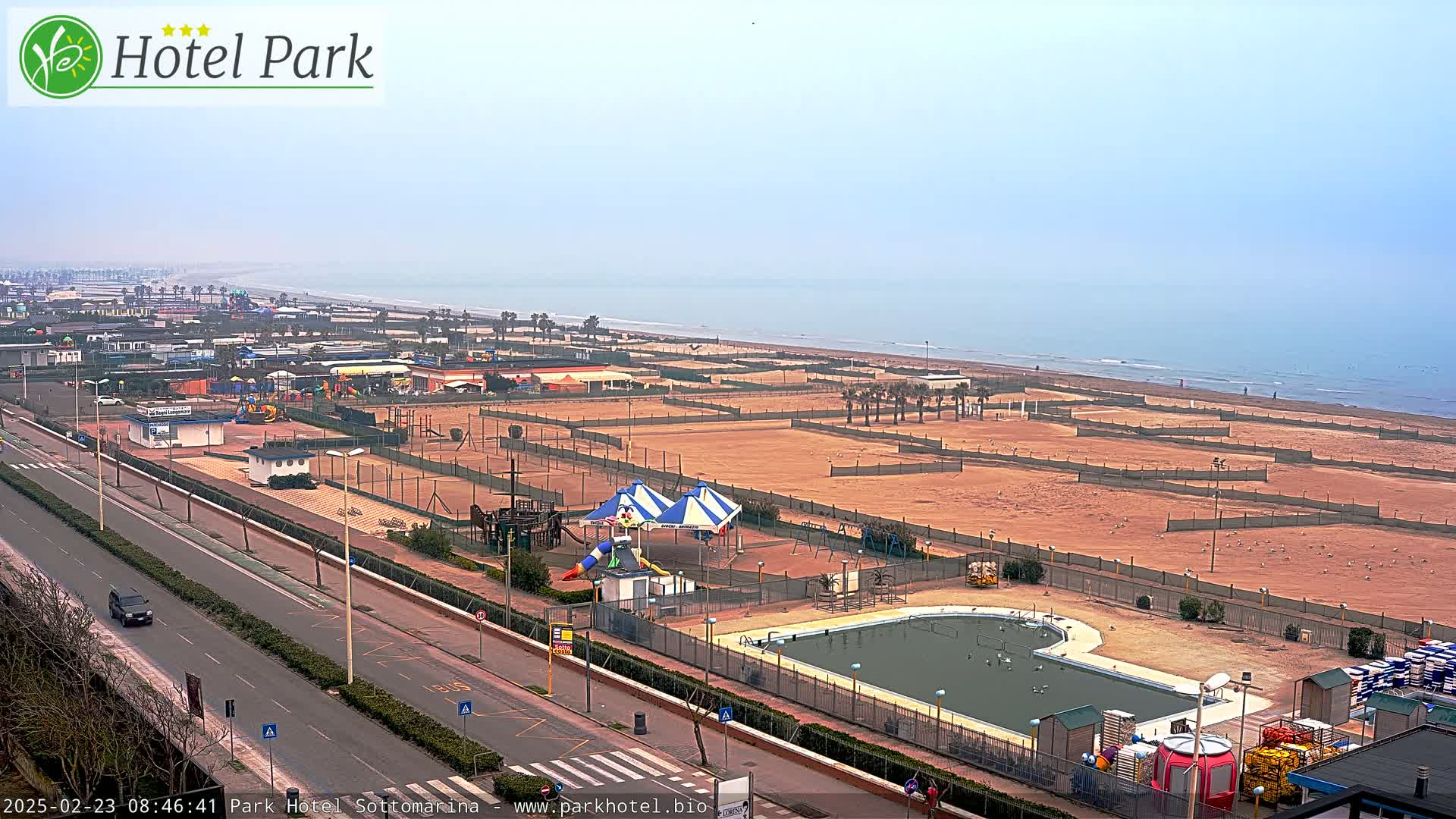 An aerial view shows a beach with a hazy sky, a road, and playground equipment near a swimming pool.