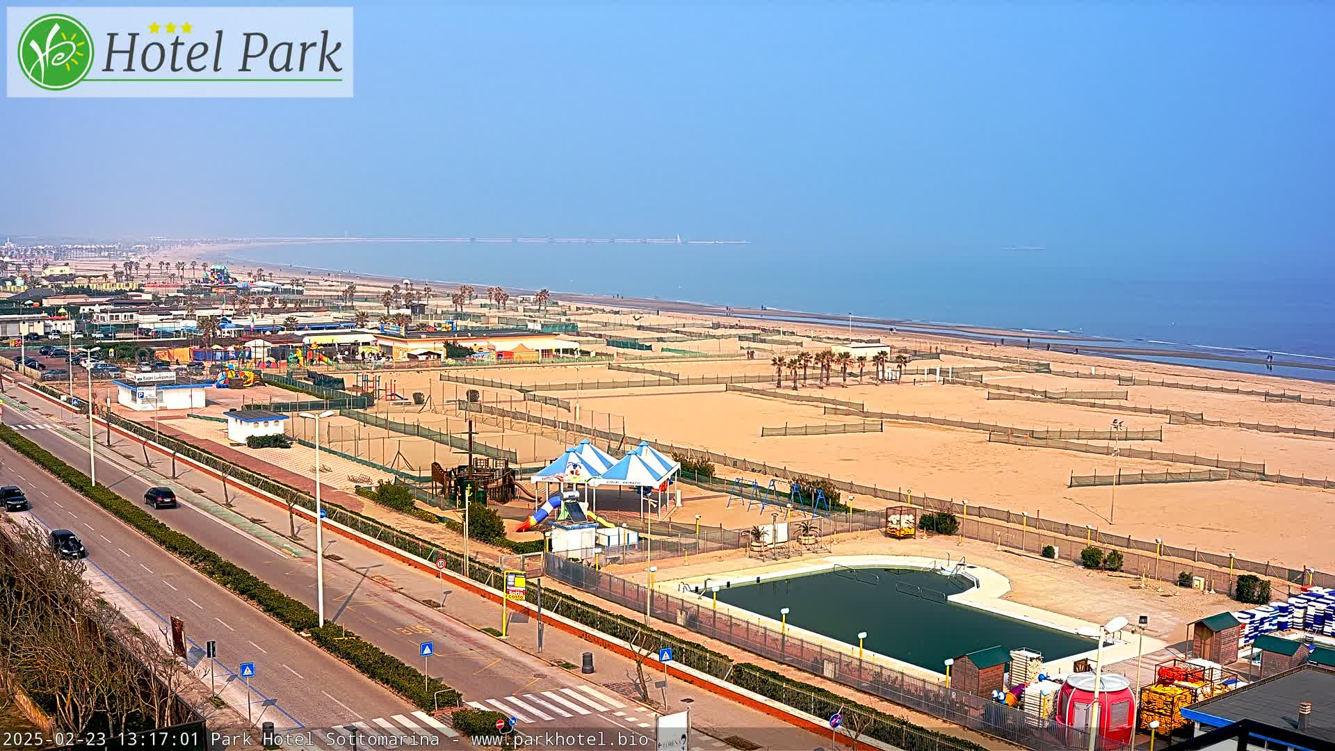 A beachfront area with a playground, swimming pool, and volleyball courts under a clear, sunny sky.