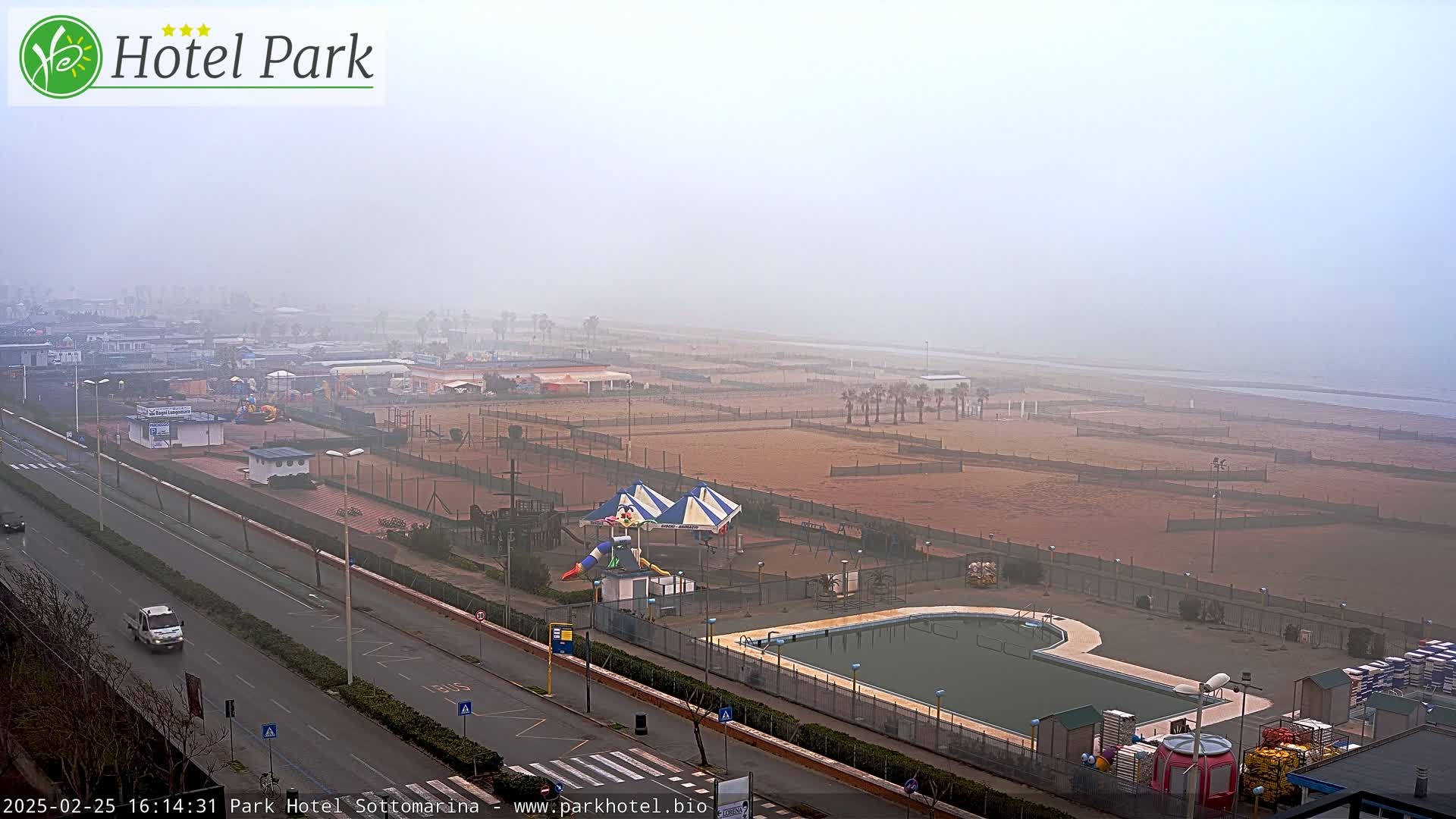 A foggy beach scene shows a resort area with a pool, playground equipment, and volleyball courts, all adjacent to a wide, sandy beach and a road.